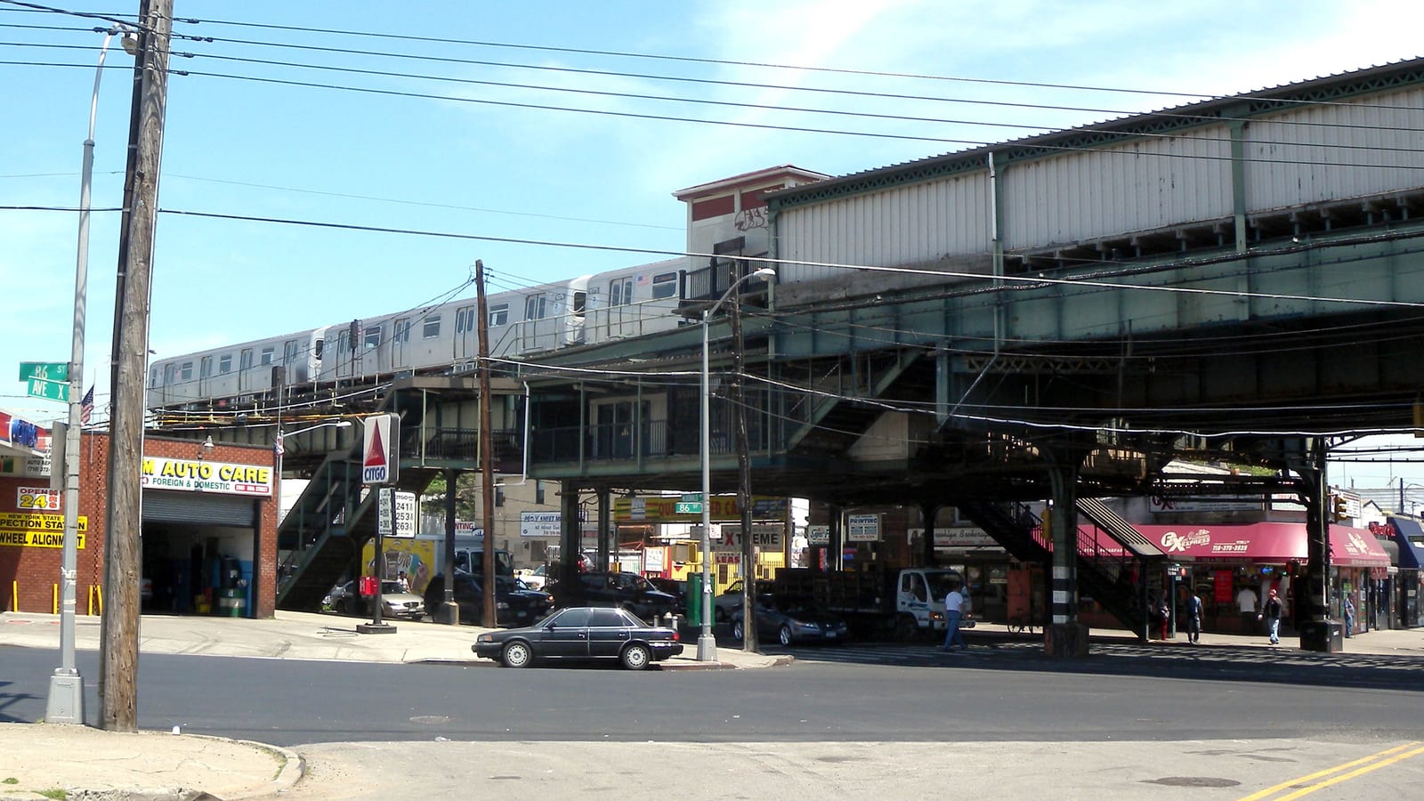 A silver subway train on an elevated steel structure over a street corner, with street signs reading 86 St and Ave X, an auto repair shop and a gas station sign at left, and shops with red awnings under the tracks at right