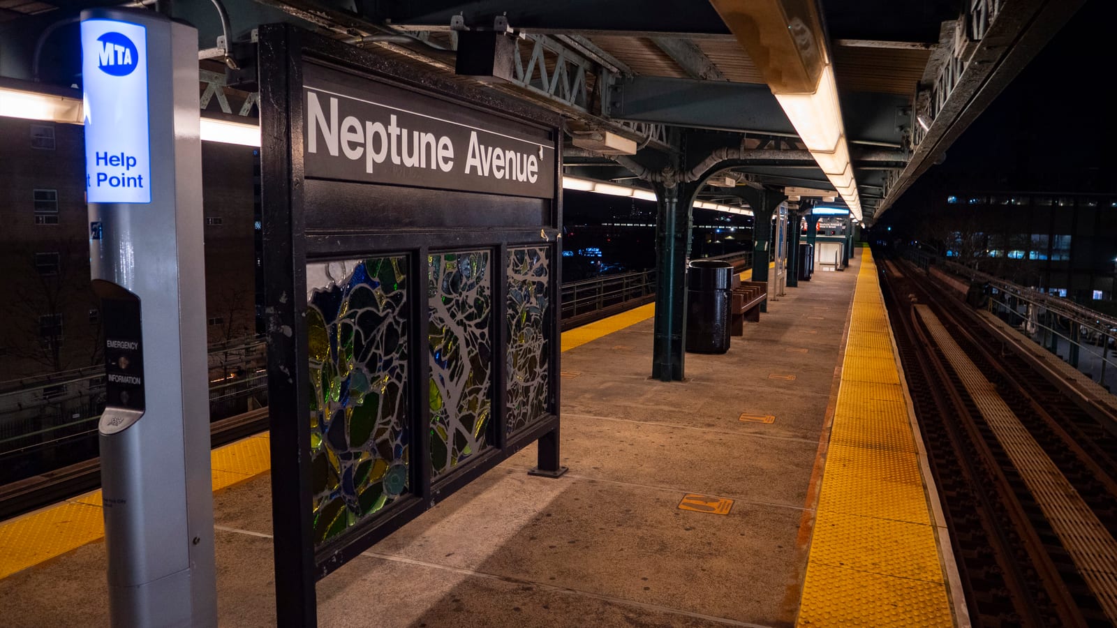 An elevated subway platform at night, with a black sign reading Neptune Avenue above stained-glass panels, a lit MTA Help Point column at left, a yellow platform edge strip and tracks at right
