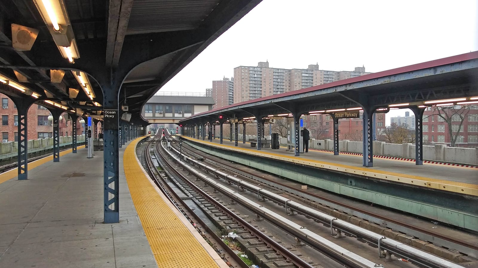 An open elevated subway station with two covered platforms on blue steel columns, signs reading Ocean Pkwy and Ocean Parkway, tracks curving between them and tall brick apartment towers beyond under a grey sky
