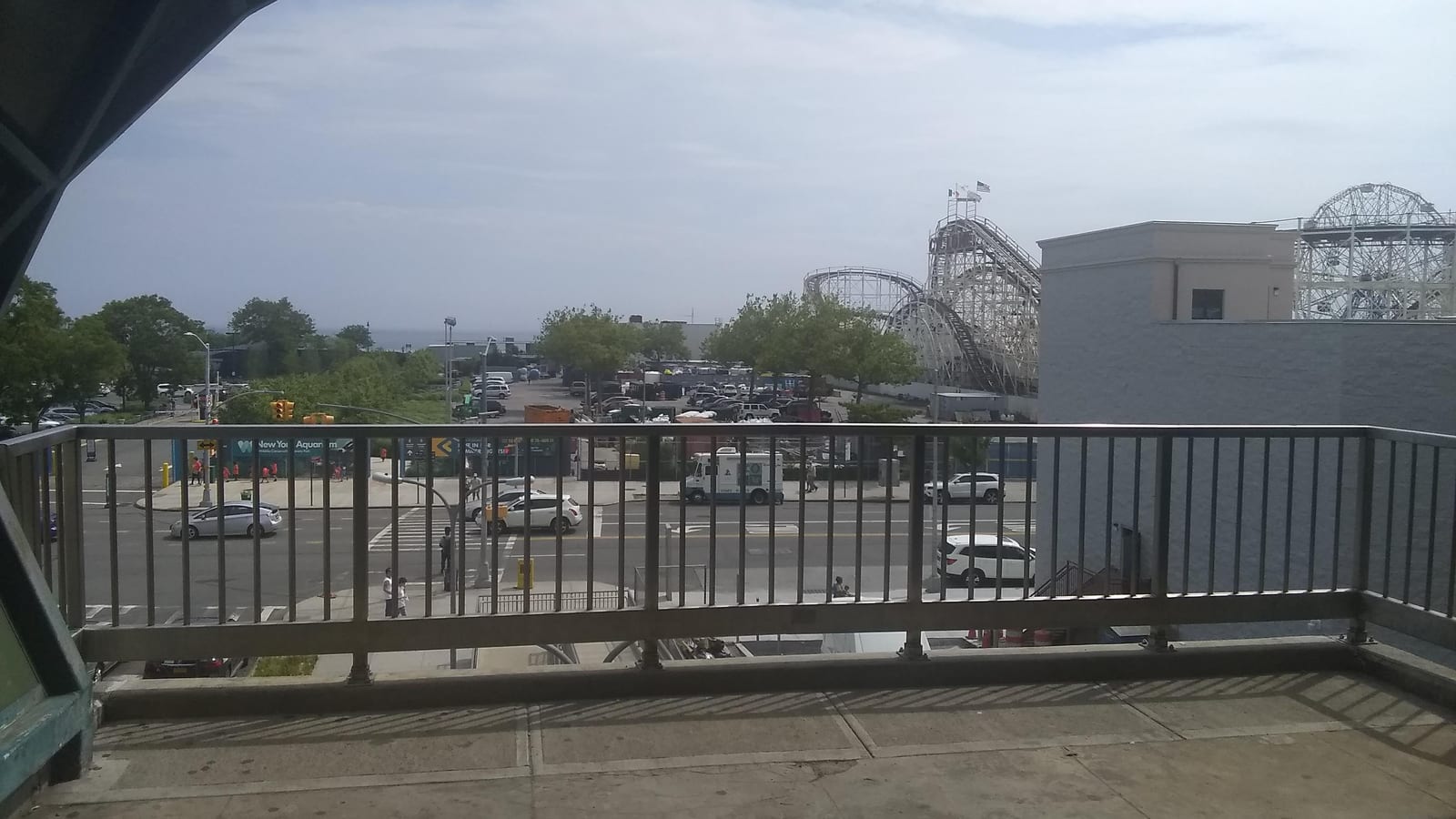 A view from an elevated station landing through a metal railing over a wide street, with parked cars, an ice cream truck, a New York Aquarium sign, trees, a wooden roller coaster and a Ferris wheel behind a pale building, and the sea on the horizon