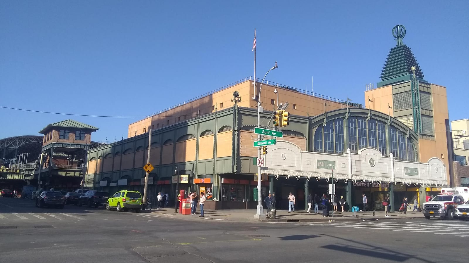 A large tan and green station building on a street corner, with arched windows over an entrance canopy marked BMT Lines, a green tower with a round ornament on top, street signs reading Surf Av and Stillwell Av, a yellow-green taxi, an ambulance and people on the sidewalk under a clear blue sky