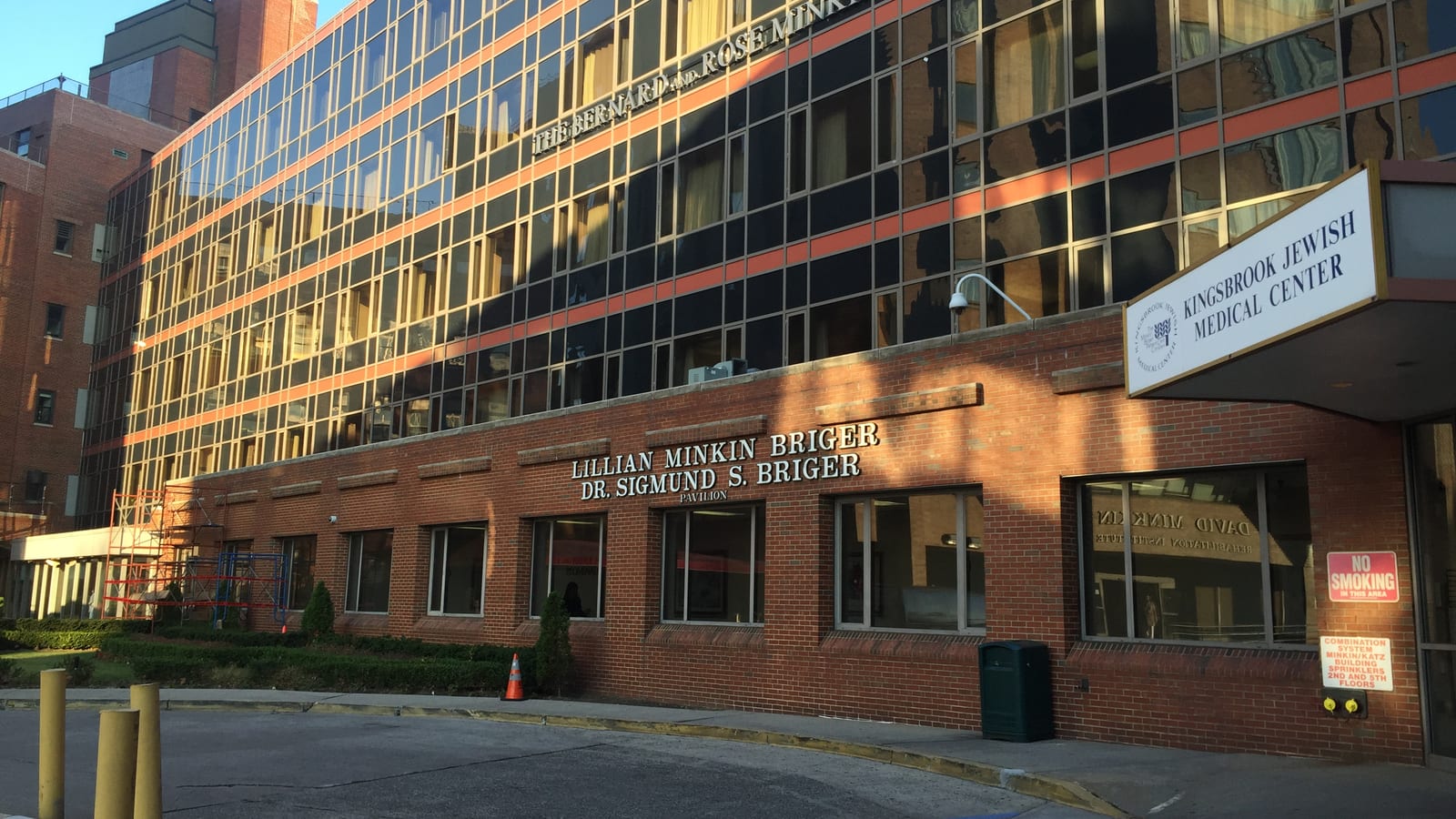 Photograph of a hospital building with a five-story glass and orange-banded upper part over a red brick ground floor lettered Lillian Minkin Briger, Dr. Sigmund S. Briger Pavilion, a canopy sign reading Kingsbrook Jewish Medical Center at right, and a curved driveway with yellow posts in the foreground