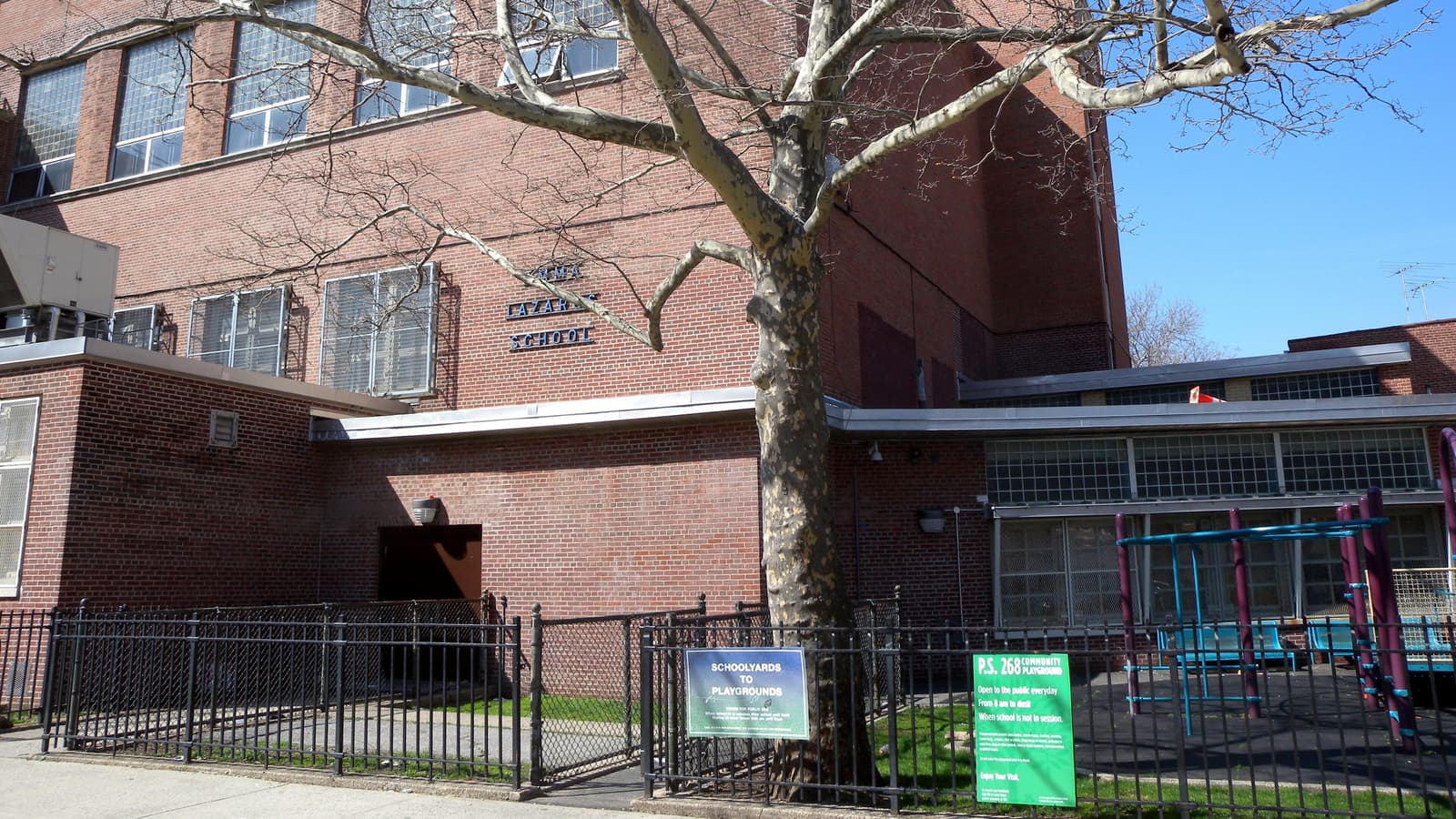 Photograph of a red brick school building with metal letters reading Emma Lazarus School, a tall bare plane tree in front, a black iron fence with a sign reading Schoolyards to Playgrounds and a green sign for the P.S. 268 community playground, and play equipment at right under a blue sky