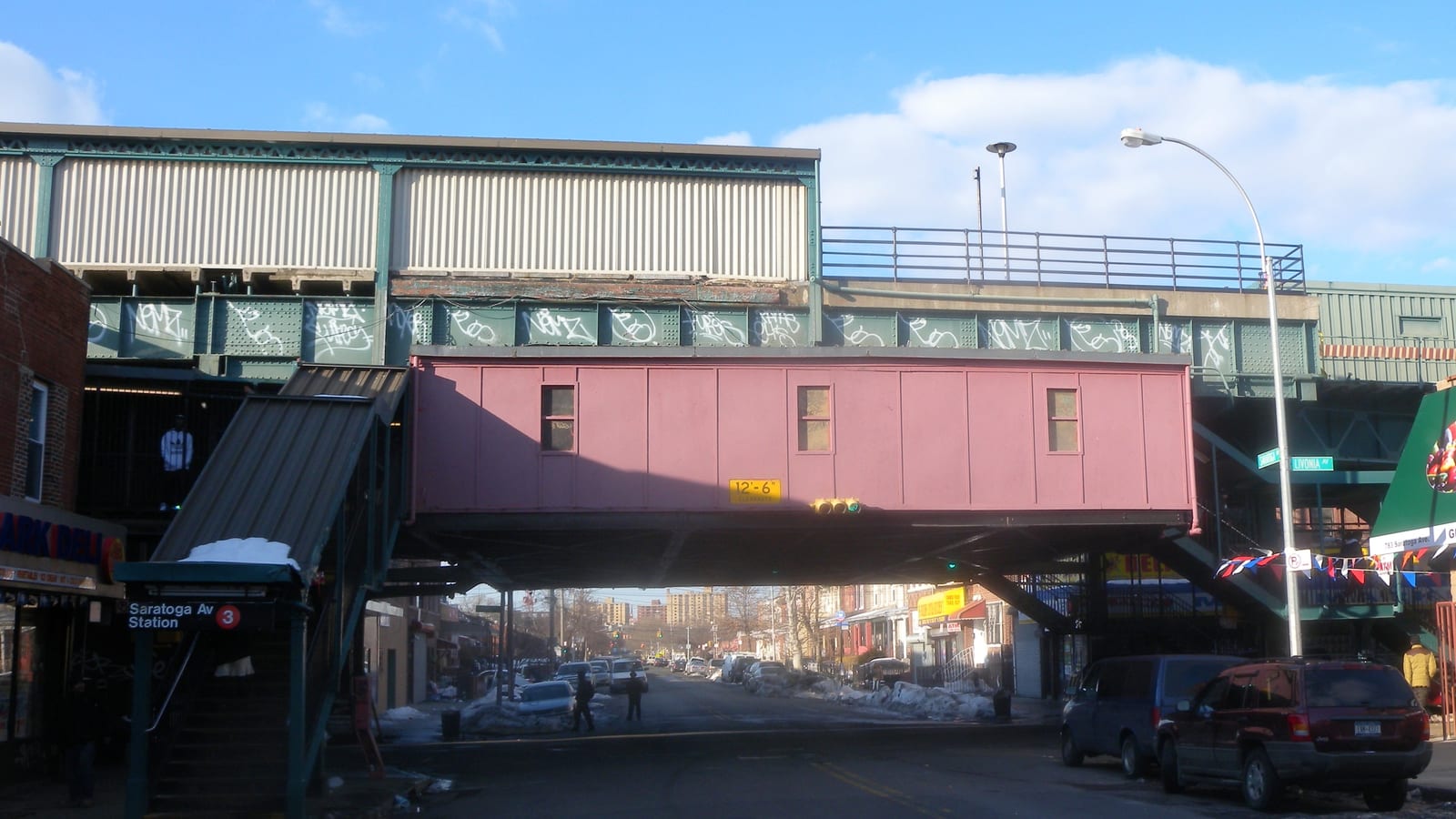 Photograph of a raised subway line crossing a street, with a pink station house marked 12 feet 6 inches, graffiti on the green steel, a covered stair at left with a sign reading Saratoga Av Station and the 3 train symbol, street signs for Livonia Avenue, parked cars, snow banks and small distant figures under a blue sky