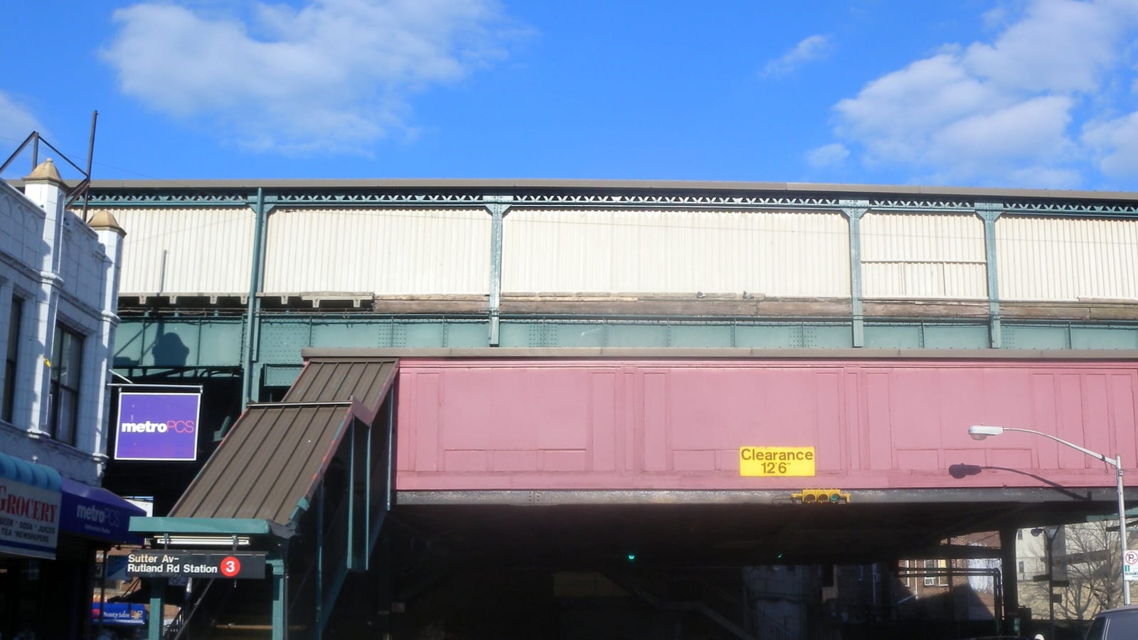 Photograph of a subway line running above a street on green steel supports, with a pink station house over the road marked Clearance 12 feet 6 inches, a covered stair at left with a sign reading Sutter Av-Rutland Rd Station and the 3 train symbol, and shops at the street corner under a blue sky