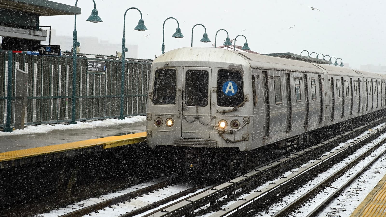 Photograph of a silver A train with its headlights on arriving at an open-air platform in falling snow, with snow on the tracks, green lamp posts and a fence with a Beach 67 Street sign at left