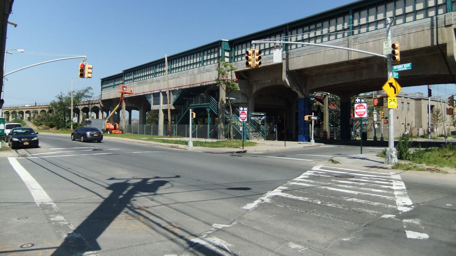 Photograph of a long elevated concrete subway station with green trim over a street corner, with stairs up to it, traffic lights, a Do Not Enter sign, a yellow curve sign and two parked cars, under a clear sky