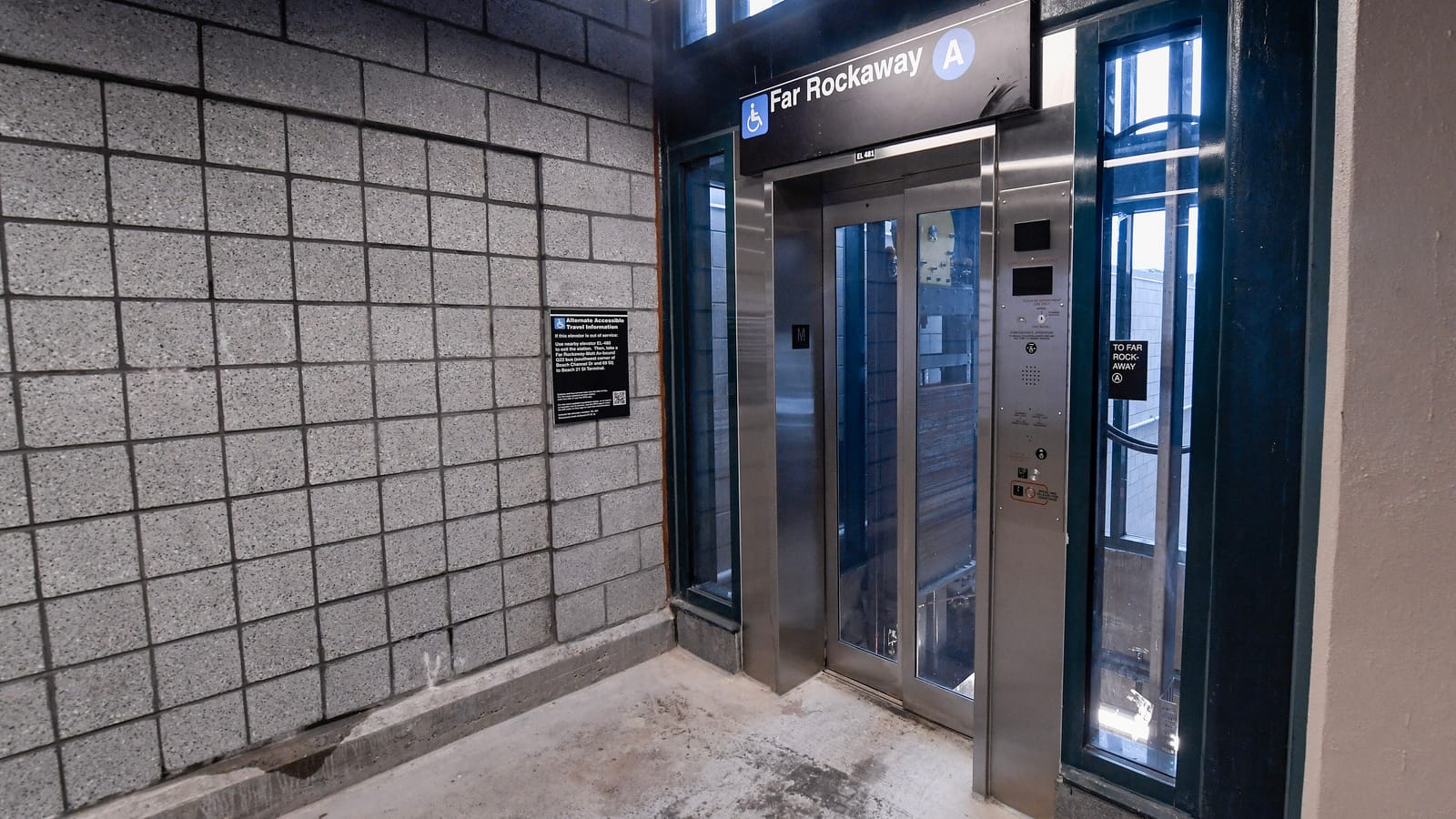 Photograph of a stainless steel elevator with glass doors under a sign reading Far Rockaway with a wheelchair symbol and an A train symbol, beside a gray block wall with a black notice about alternate accessible travel