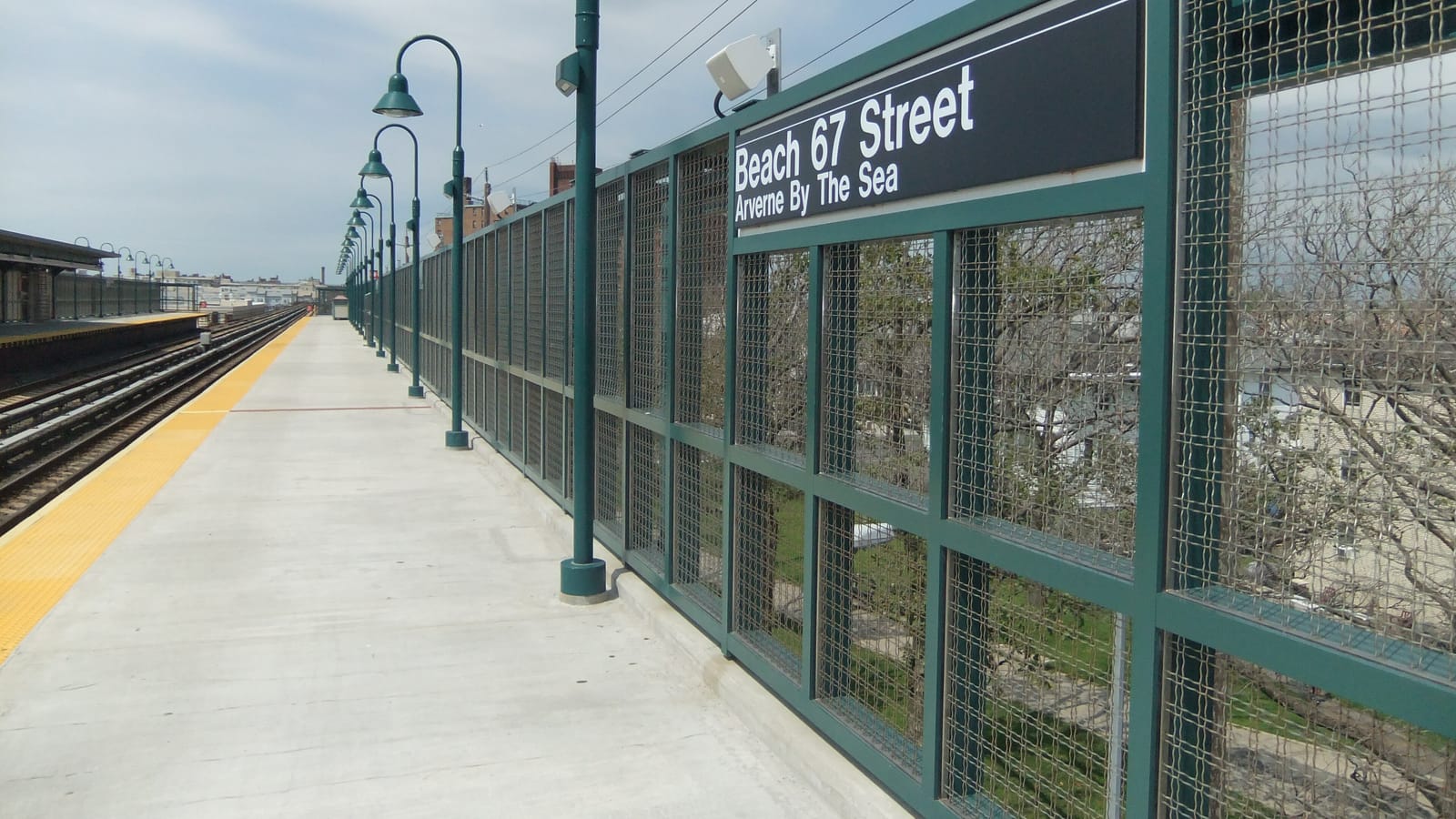 Photograph of an empty open-air subway platform with a yellow edge strip, green lamp posts, a green mesh fence at right with a black sign reading Beach 67 Street, Arverne By The Sea, and trees and houses beyond the fence