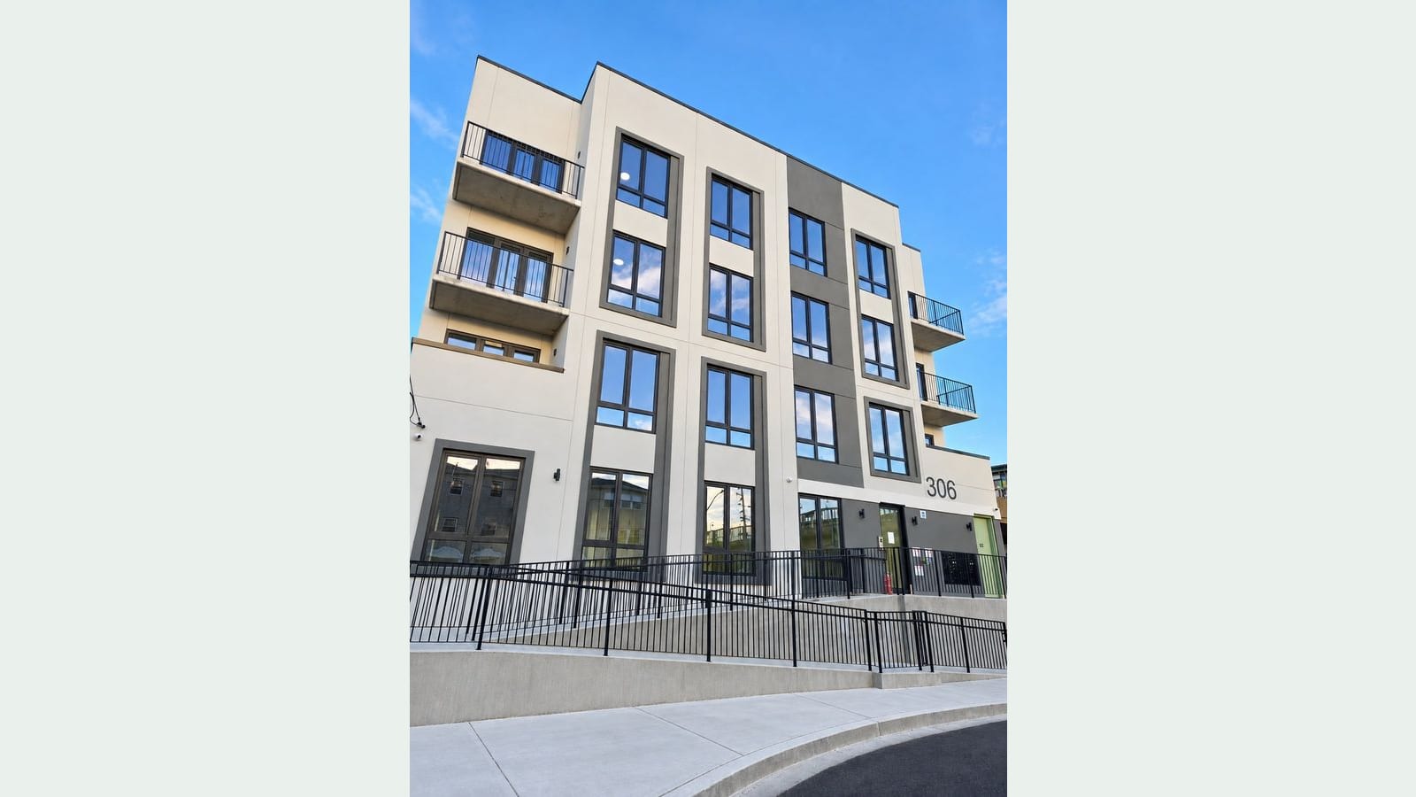 Photograph looking up at a new 4-story building with a white and dark gray facade, tall dark-framed windows, balconies with black railings at both ends, the number 306 beside the entrance, a ramp and stairs with black railings in front, and a curved sidewalk under a blue sky
