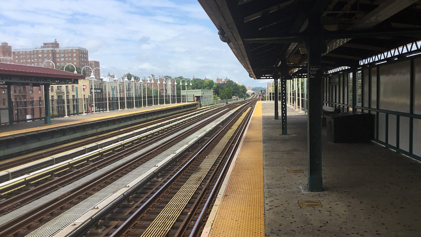 Photograph from an elevated subway platform looking along three sets of tracks under a blue sky with clouds, with a yellow platform edge, a covered platform and green columns on the right, and apartment buildings and lamp posts in the distance