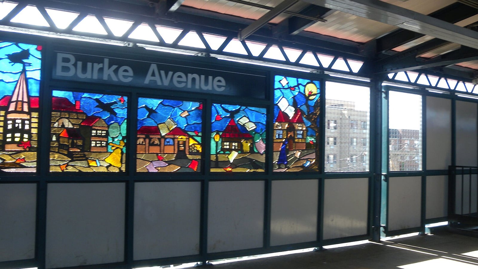 Photograph of an elevated subway platform windbreak with a row of colorful stained-glass panels showing houses, roofs and a church steeple, below a black sign reading Burke Avenue, with sunlight casting colored patches on the platform floor