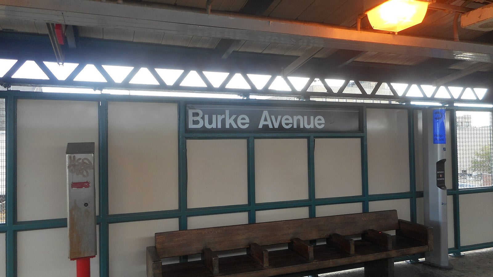 Photograph of an elevated subway platform with a gray sign reading Burke Avenue on a white panel wall with green steel framing, a wooden bench below the sign, a red-based help point on the left, an orange ceiling light above and apartment buildings visible through the gaps in the wall