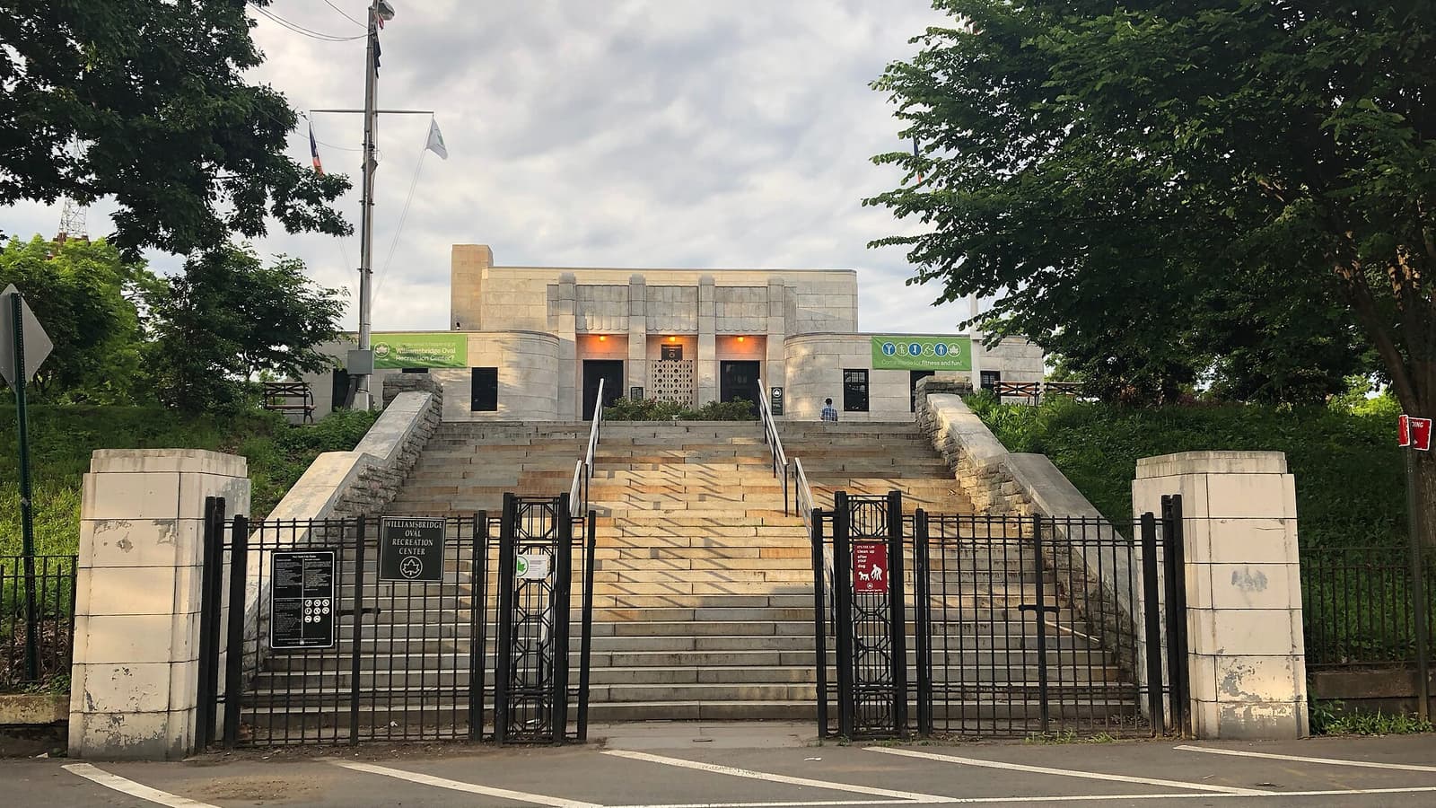 Photograph of a wide stone staircase between two square stone gateposts and black iron gates, leading up to a pale stone recreation building with green signs, framed by leafy trees under a cloudy sky, with a crosswalk in the foreground