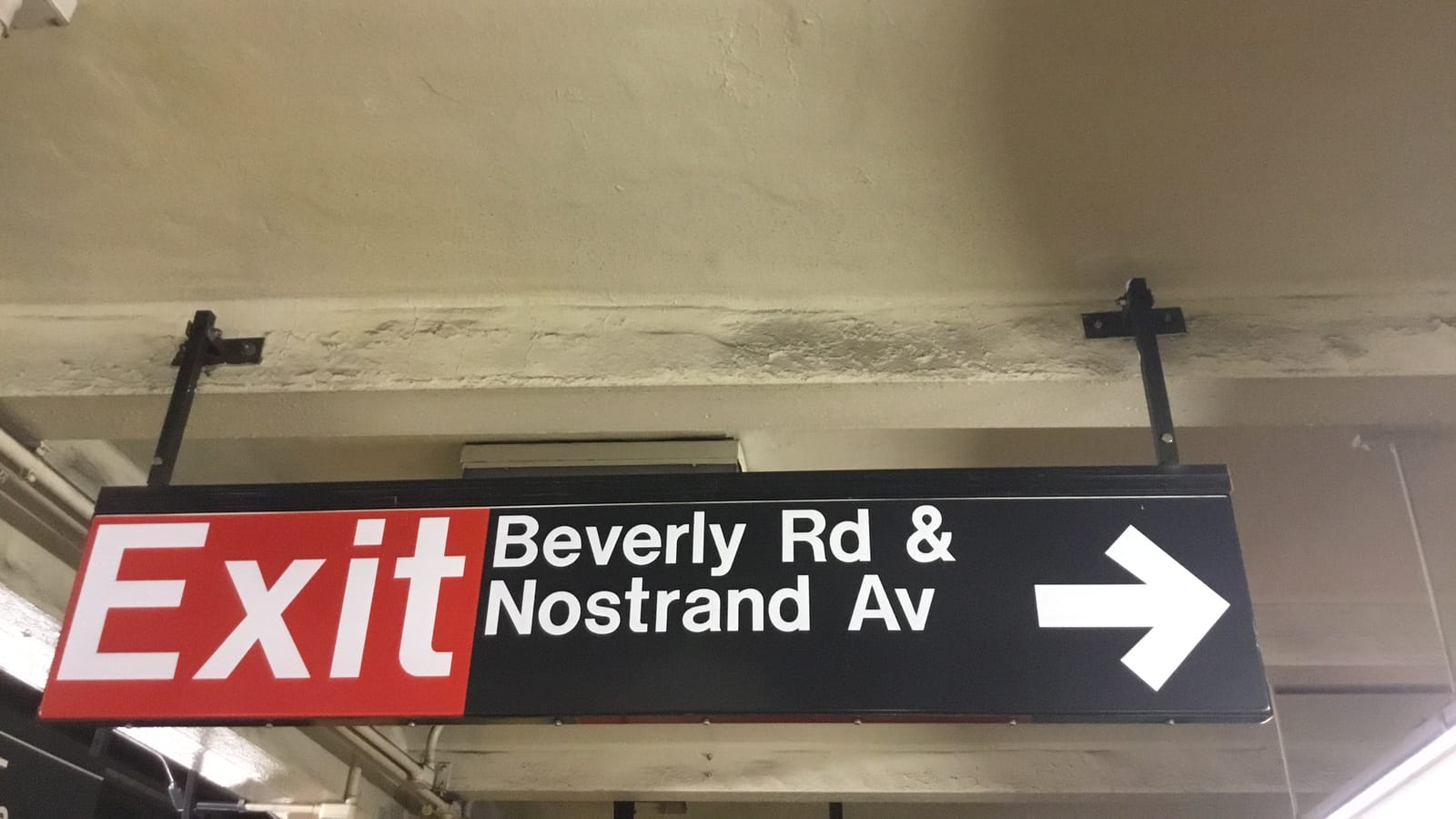 Photograph of a black hanging subway sign with a red panel reading Exit and white text reading Beverly Rd & Nostrand Av beside an arrow pointing right, hung from a stained concrete ceiling
