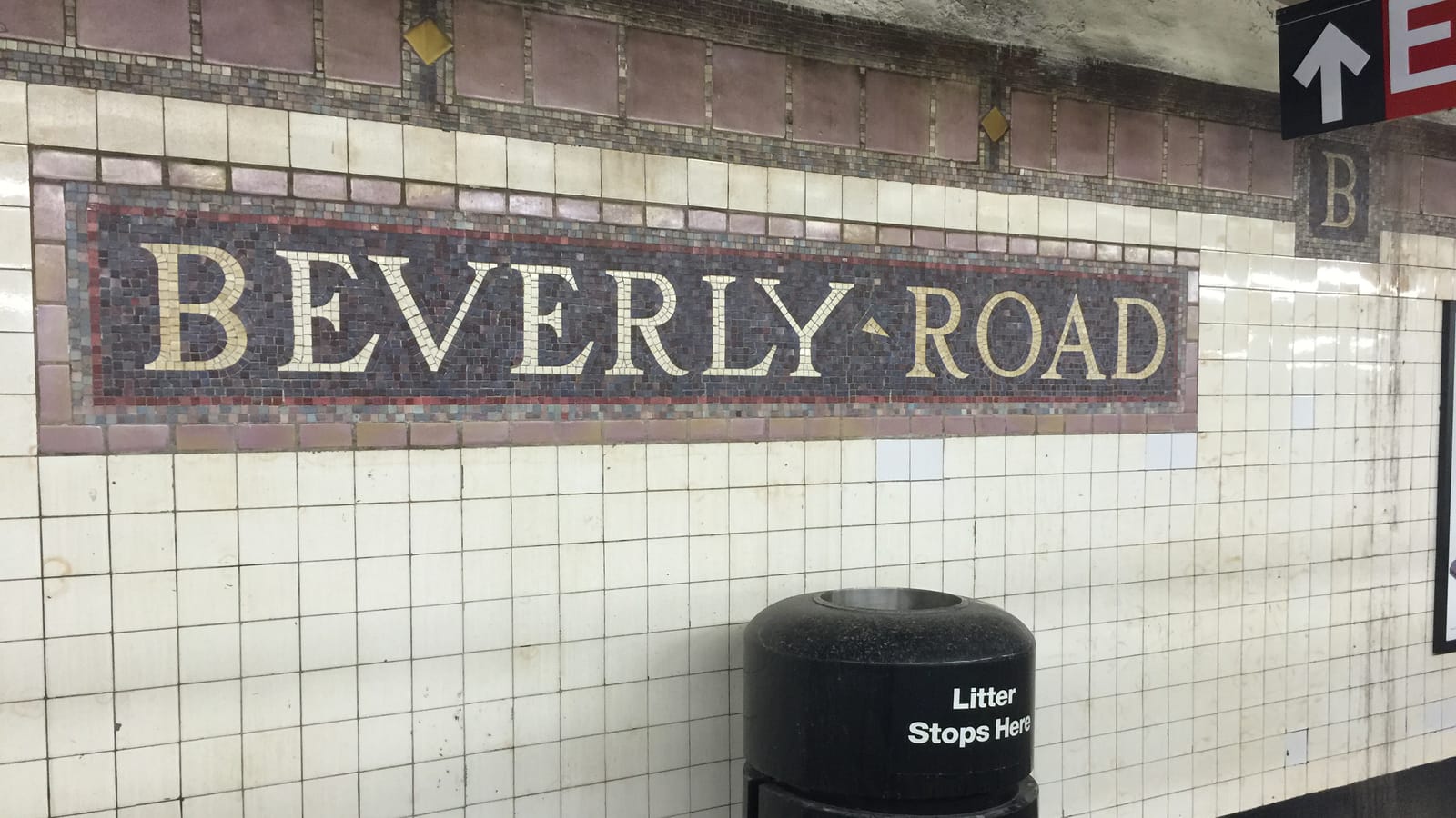 Photograph of a subway station wall with a mosaic band reading Beverly Road in cream letters on dark purple tile, a small B tile at right, white square wall tiles below, a black trash can marked Litter Stops Here and the edge of an exit sign at top right