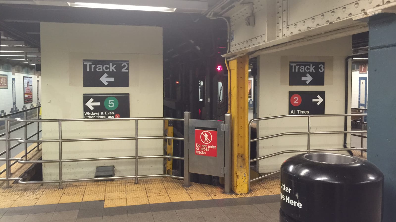 Photograph of the end of a subway platform with a metal railing and a gate marked Do not enter or cross tracks, signs reading Track 2 with a 5 train symbol for weekdays and evenings, and Track 3 with a 2 train symbol for all times, and a train's lights in the dark between the walls