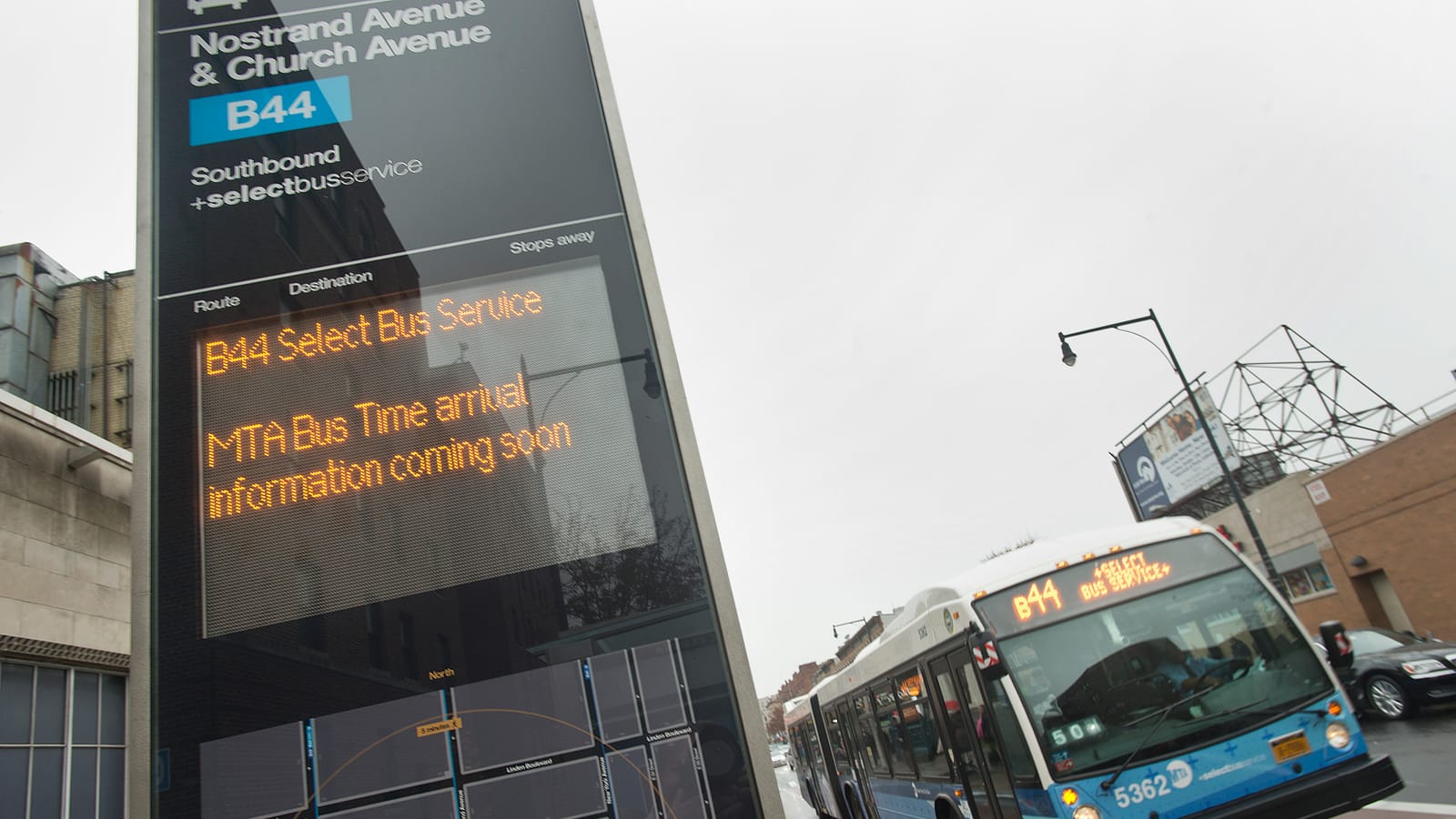 Photograph of a tall black bus stop sign reading Nostrand Avenue & Church Avenue, B44 Southbound, Select Bus Service, with an orange text display reading B44 Select Bus Service, MTA Bus Time arrival information coming soon, and a blue and white articulated bus marked B44 Select Bus Service on the street behind it under a gray sky