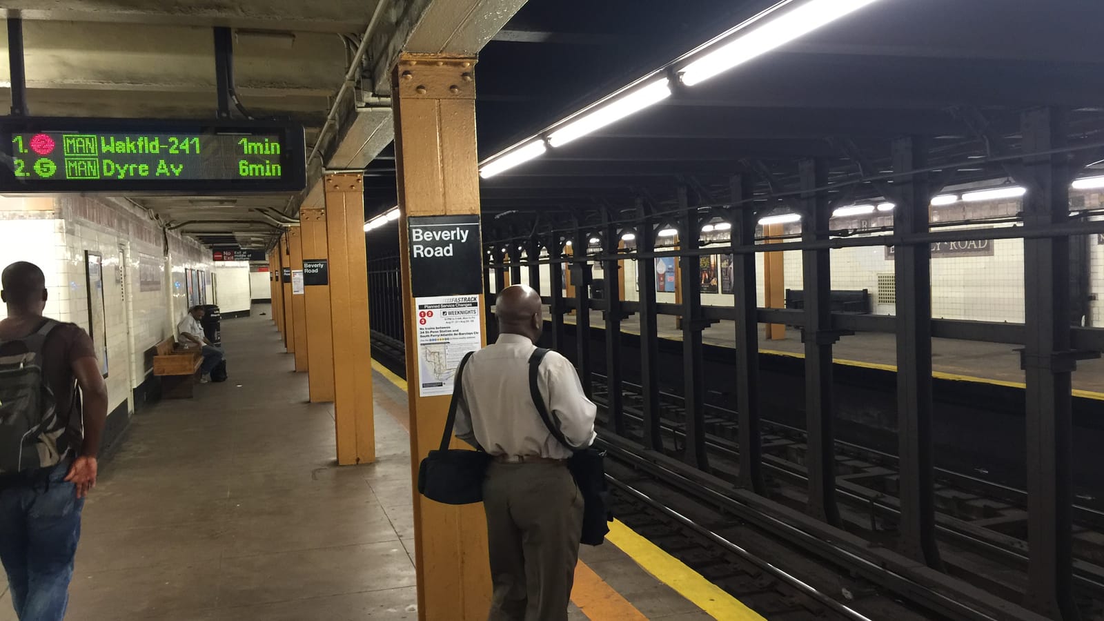 Photograph of an underground subway platform with yellow columns, a black sign reading Beverly Road, a countdown board showing a 2 train to Wakefield-241 in 1 minute and a 5 train to Dyre Av in 6 minutes, two men seen from behind and a man seated on a bench in the distance