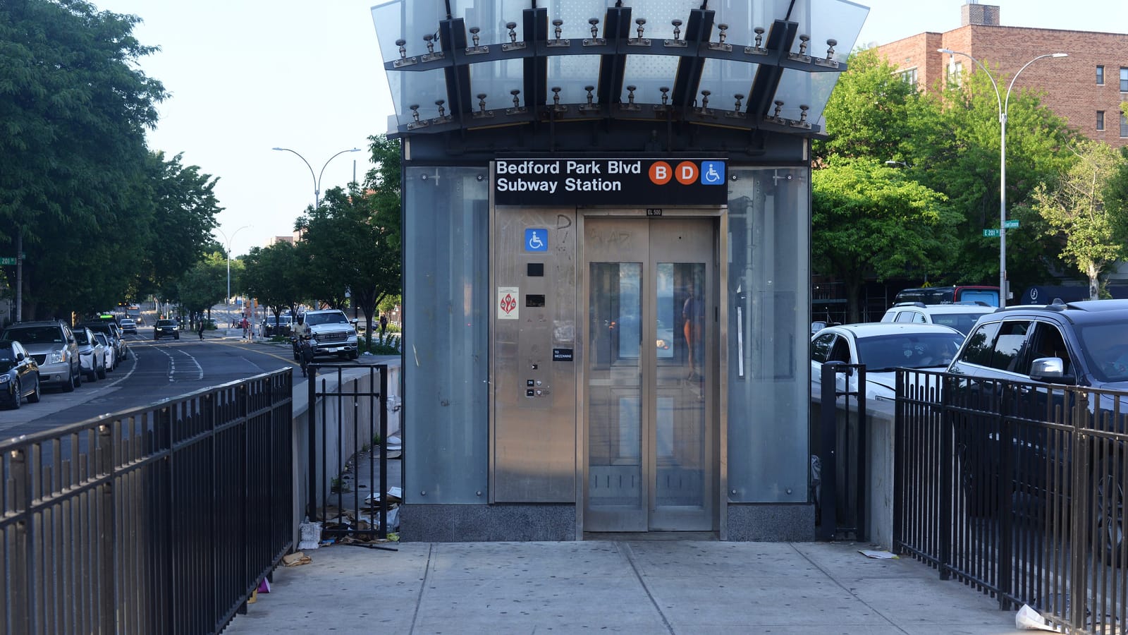 A glass and steel street elevator for the subway under a curved glass canopy, signed Bedford Park Blvd Subway Station with orange B and D symbols and a wheelchair symbol, behind black metal railings, with a wide street of parked cars and trees at left and a brick apartment building at right