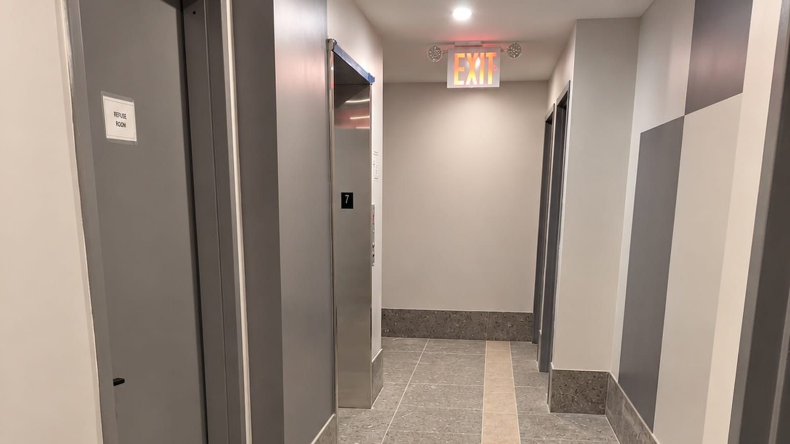 A building hallway with grey terrazzo-style floor tiles and a tan stripe down the middle, grey door frames, a door marked Refuse Room at the left, a steel elevator door with a black sign reading 7 beside it, and a lit red EXIT sign on the far wall