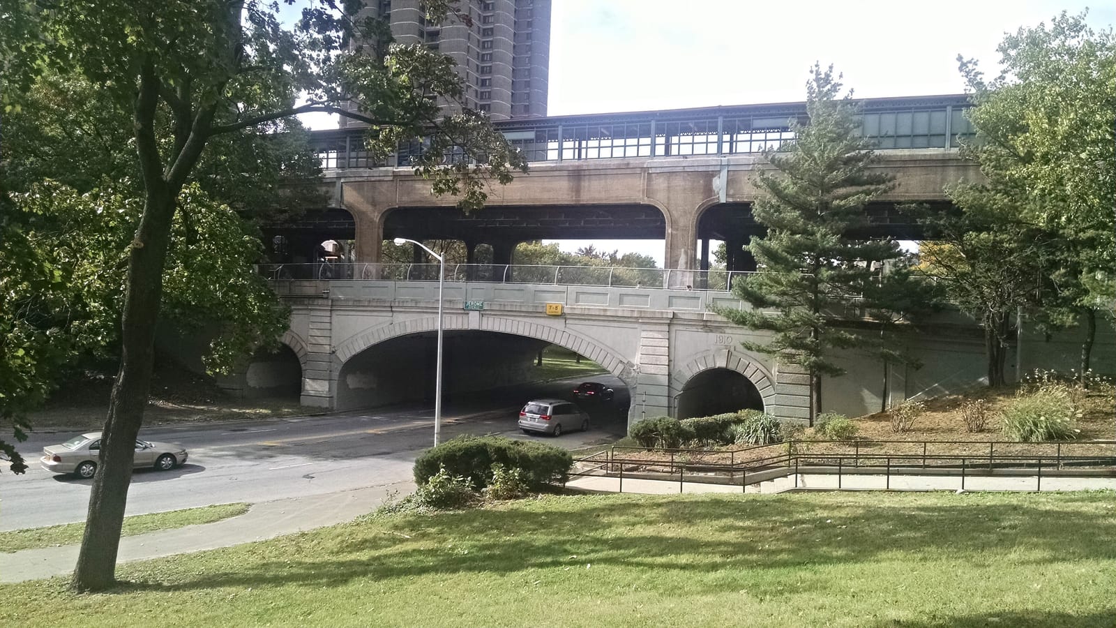 An elevated subway station with green-framed windows on a concrete viaduct, above a stone arch bridge marked Jerome and 1910 over a road with two cars passing under it, with trees, a lawn and a tall apartment tower behind