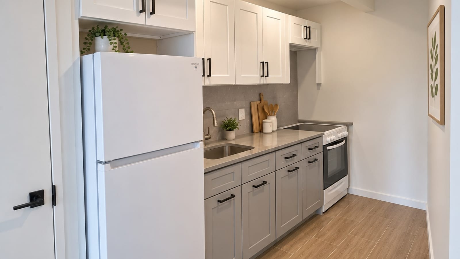 A narrow galley kitchen with white upper cabinets and grey lower cabinets with black handles, a grey stone counter with a steel sink and a curved faucet, a white refrigerator at the left, a white electric range with a smooth black cooktop at the far end, wood-look plank flooring, and potted plants, cutting boards and a framed leaf print placed for the photo