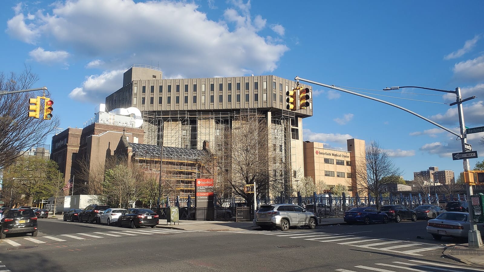 Photograph of a street corner with traffic lights, a large concrete hospital building partly covered in scaffolding, an older brick building beside it, a tan building with a sign reading Interfaith Medical Center, parked cars along the curb and a Herkimer street sign at right under a blue sky with clouds