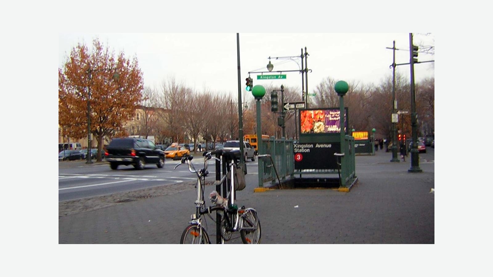 Photograph of a subway entrance with green railings and two green globe lamps, a sign reading Kingston Avenue Station with a 3 bullet, a folding bicycle locked to a post in front, a wide avenue with traffic and a yellow taxi at left, autumn trees and a green Kingston Av street sign, shown at its original size on a plain light background