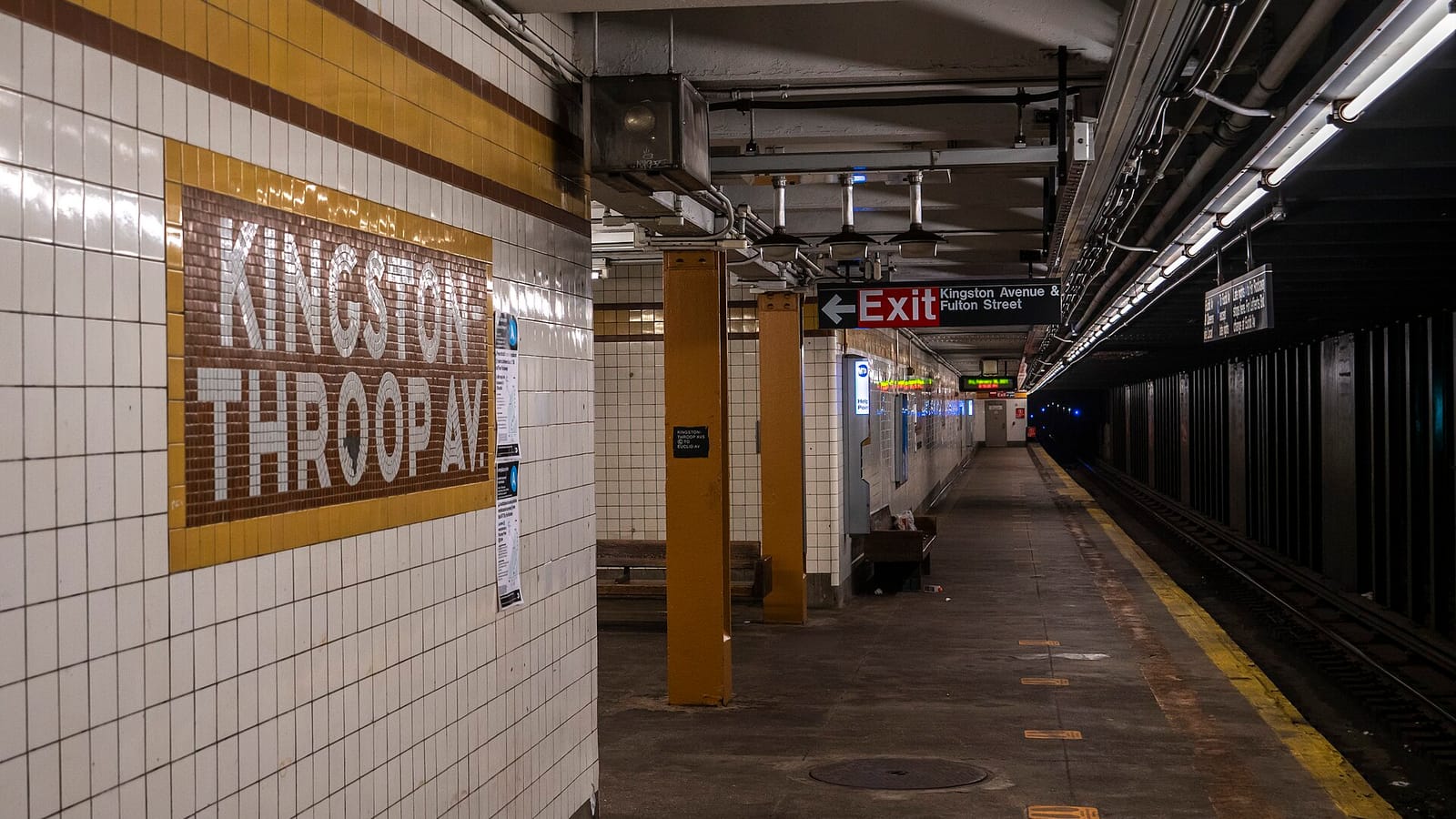Photograph of an underground subway platform with white tile walls, a brown and white mosaic reading Kingston-Throop Av., mustard-colored columns, an Exit sign pointing to Kingston Avenue and Fulton Street, and an empty platform beside the tracks