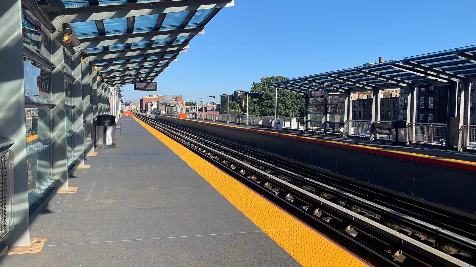 Photograph of an elevated rail platform on a sunny day, with a glass and steel canopy on the left, a yellow tactile strip along the platform edge, two tracks, and a second platform on the right with a sign reading Nostrand, brick row houses and trees beyond under a clear blue sky