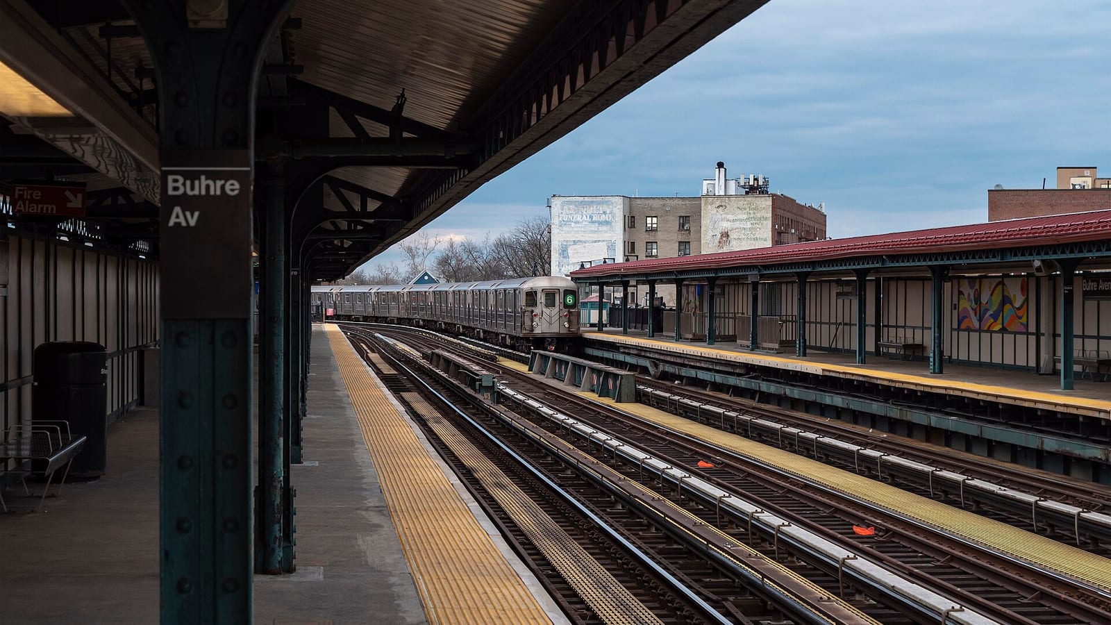 Photograph along a covered elevated subway platform with a green column marked Buhre Av, looking down three tracks toward a silver 6 train in the distance, with the opposite platform's red roof and a colorful wall mural on the right and older brick buildings behind under a blue-gray sky