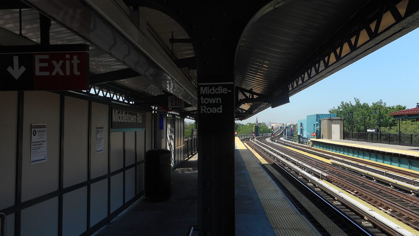 Photograph from under the roof of an elevated subway platform, with a red Exit sign, a column labelled Middle-town Road, a Middletown Rd station sign on the wall and posted service notices, looking out along curving tracks toward green trees under a clear blue sky