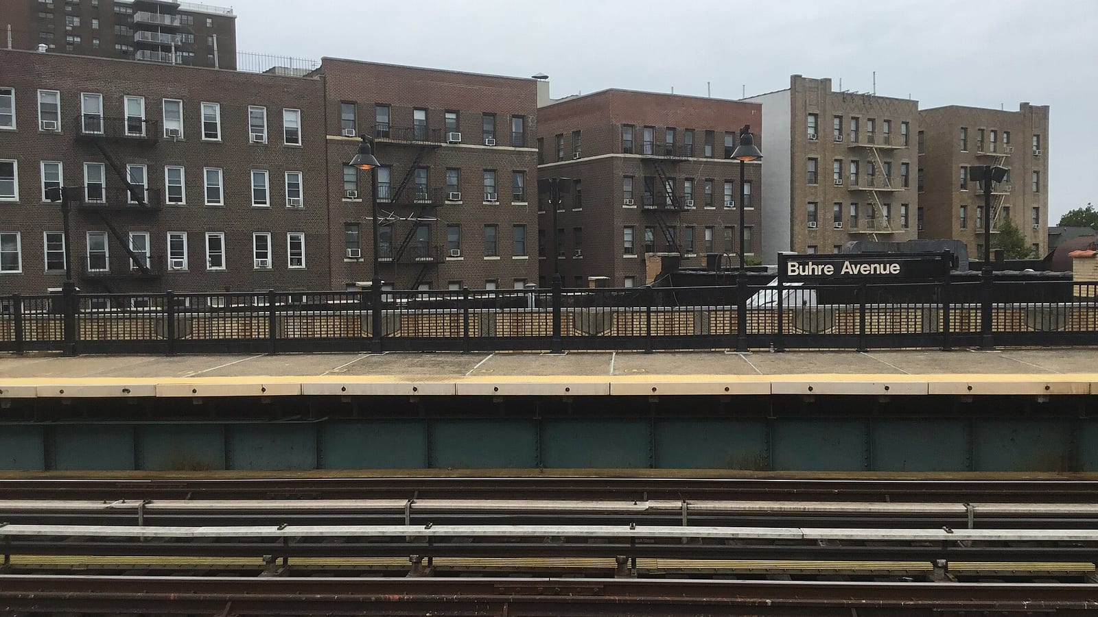 Photograph from an elevated subway platform across the tracks to the other platform, with a black railing and a sign reading Buhre Avenue, and a row of four- to six-story brick and tan apartment buildings with fire escapes and window air conditioners behind it under a gray sky