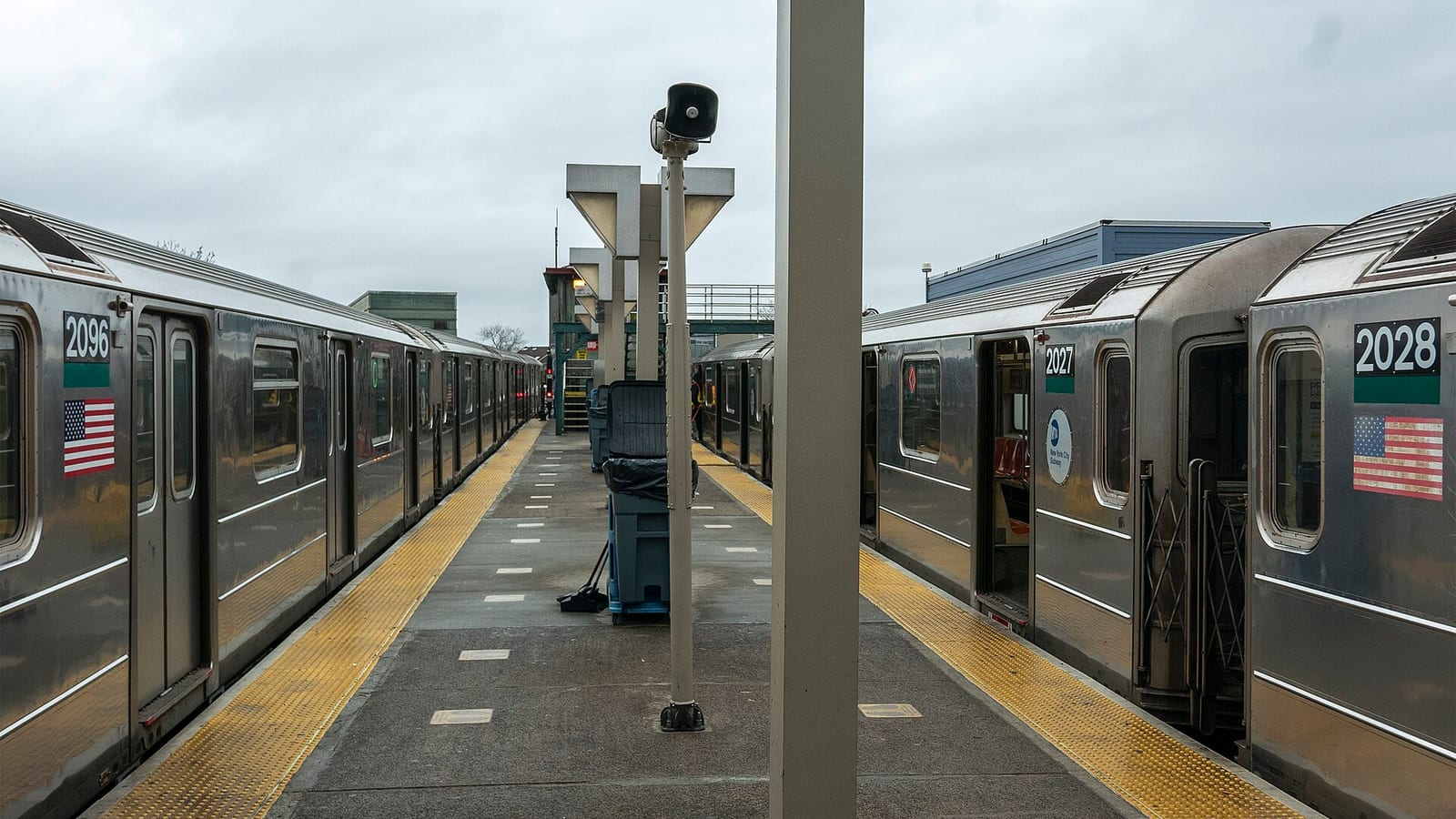 Photograph of an elevated subway platform under a cloudy sky, with two silver subway trains parked on either side, car numbers 2096, 2027 and 2028 and American flag decals, a loudspeaker pole and a column in the middle of the platform, and yellow tactile strips along both edges