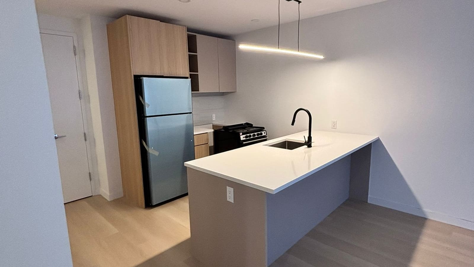 Photograph of an empty apartment kitchen with a white-topped peninsula holding a sink and a black faucet, a stainless-steel top-freezer refrigerator set in a tall wood-tone cabinet, grey upper cabinets, a black freestanding range with coil burners, a linear pendant light and pale wood-look flooring