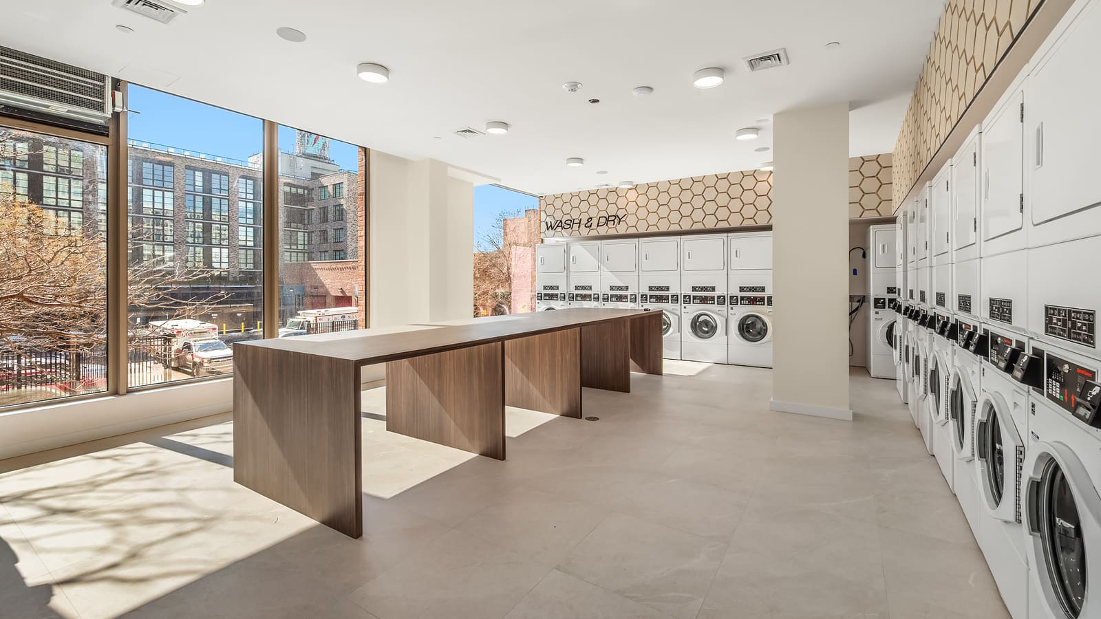 Photograph of a laundry room with rows of white front-loading washers and stacked dryers along two walls under a hexagon-patterned band and a sign reading WASH & DRY, a long wood folding table in the middle, pale floor tile and floor-to-ceiling windows looking onto a street and a grey apartment building
