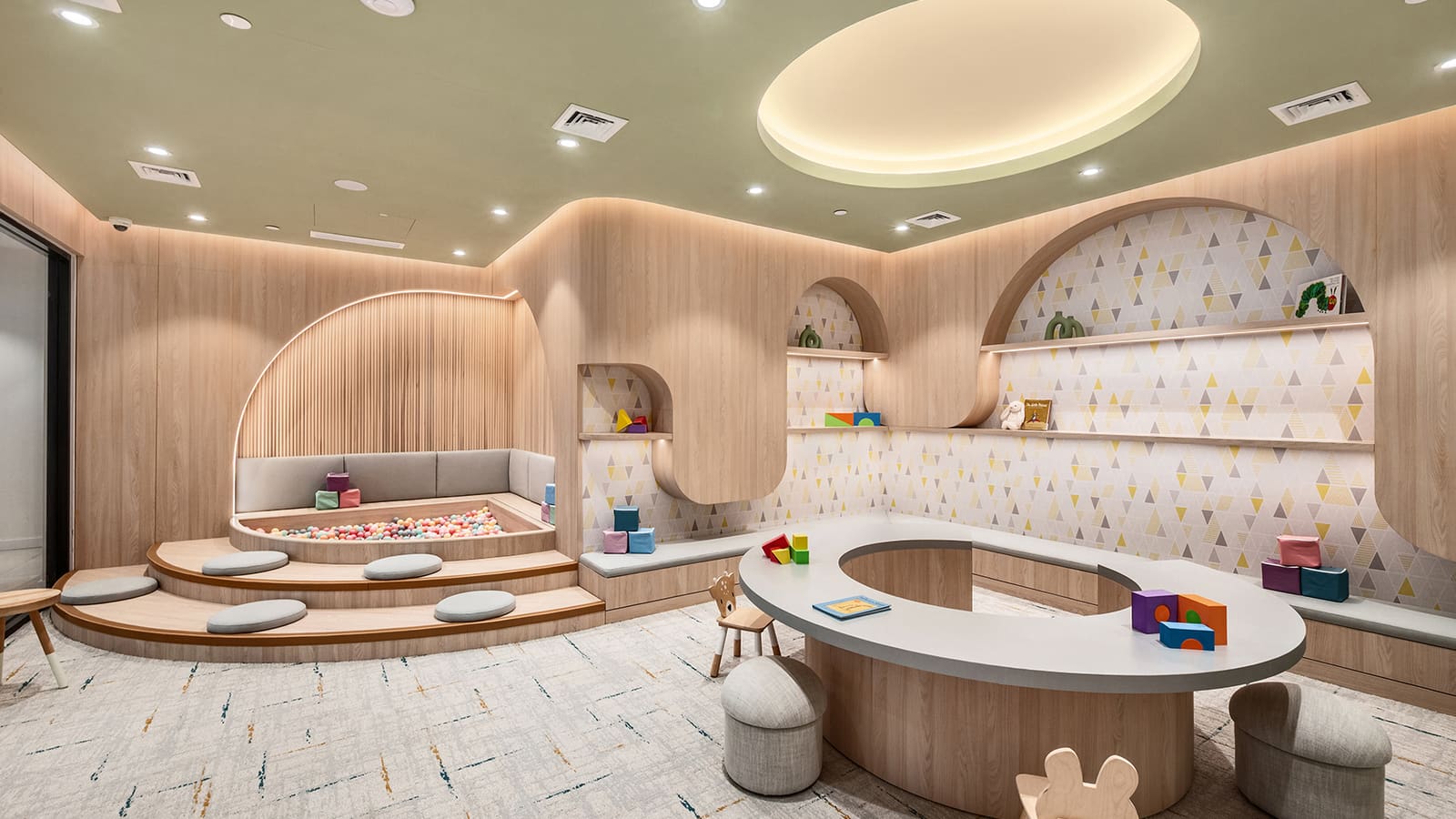 Photograph of a children's playroom with curved wood-panelled walls, a sage-green ceiling with an oval light, a ball pit set into carpeted steps with round cushions, shelves of toys and picture books against triangle-patterned wallpaper, and a ring-shaped play table with small stools