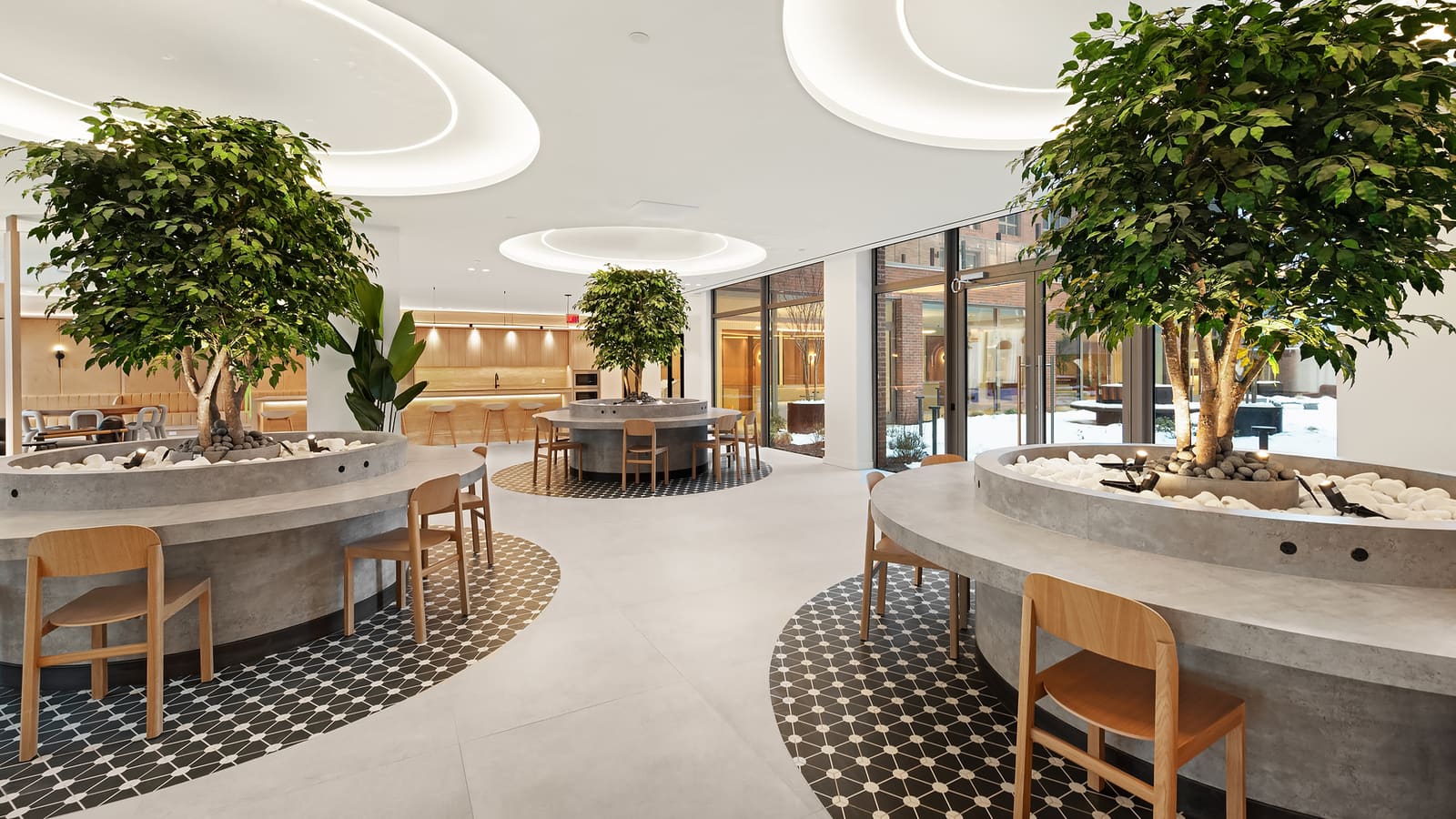 Photograph of a lounge with three round concrete planters holding indoor trees, each ringed by a counter with wooden chairs on black-and-white patterned floor tile, oval lit recesses in a white ceiling, a kitchen counter with stools in the background and glass doors onto a snow-covered courtyard with brick walls