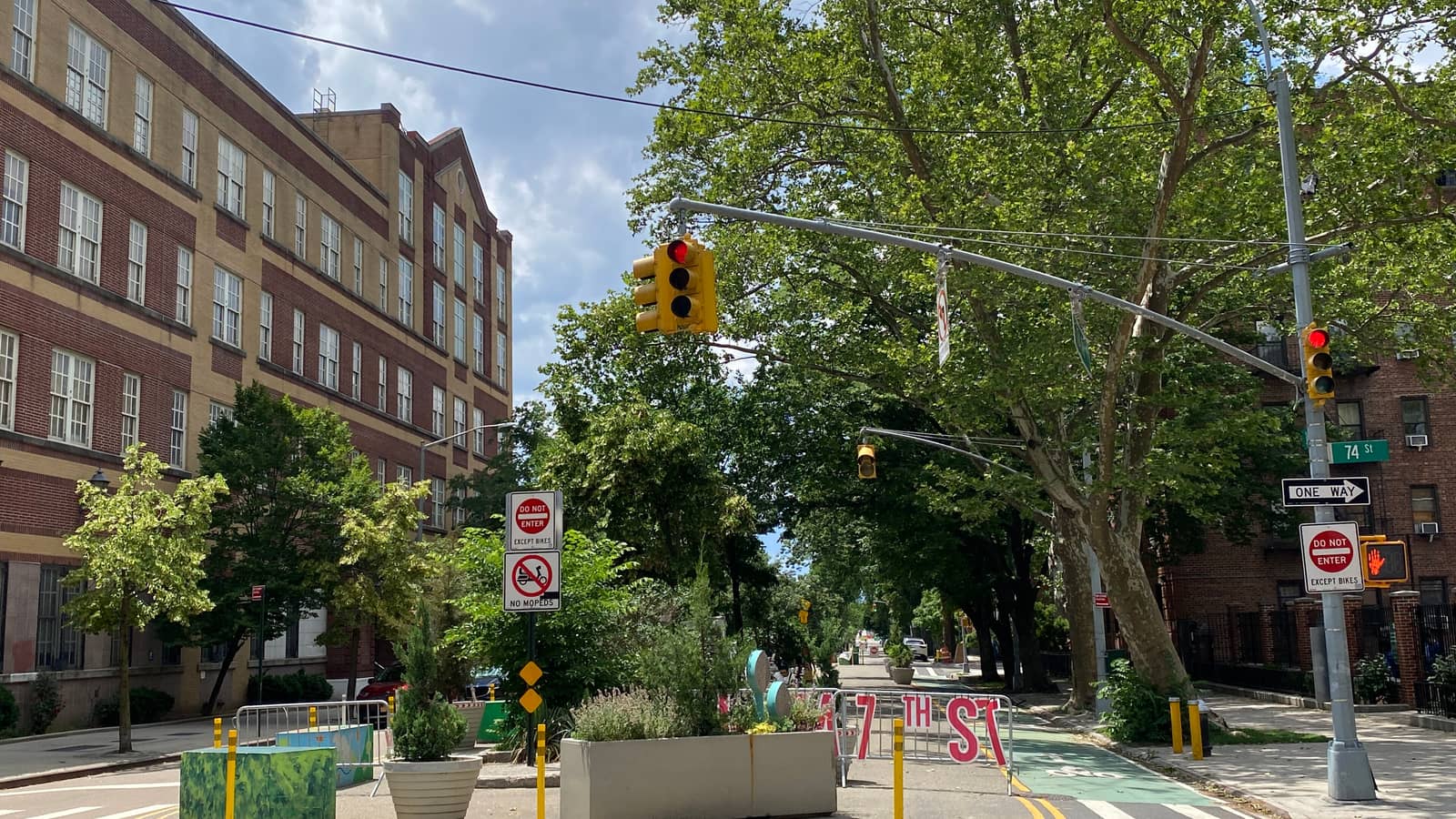 A tree-lined avenue closed to cars with planters, bollards, a painted street barrier, Do Not Enter Except Bikes signs, a green bike lane, traffic lights, a 74 St street sign, a large brick school building at left and brick apartment buildings at right