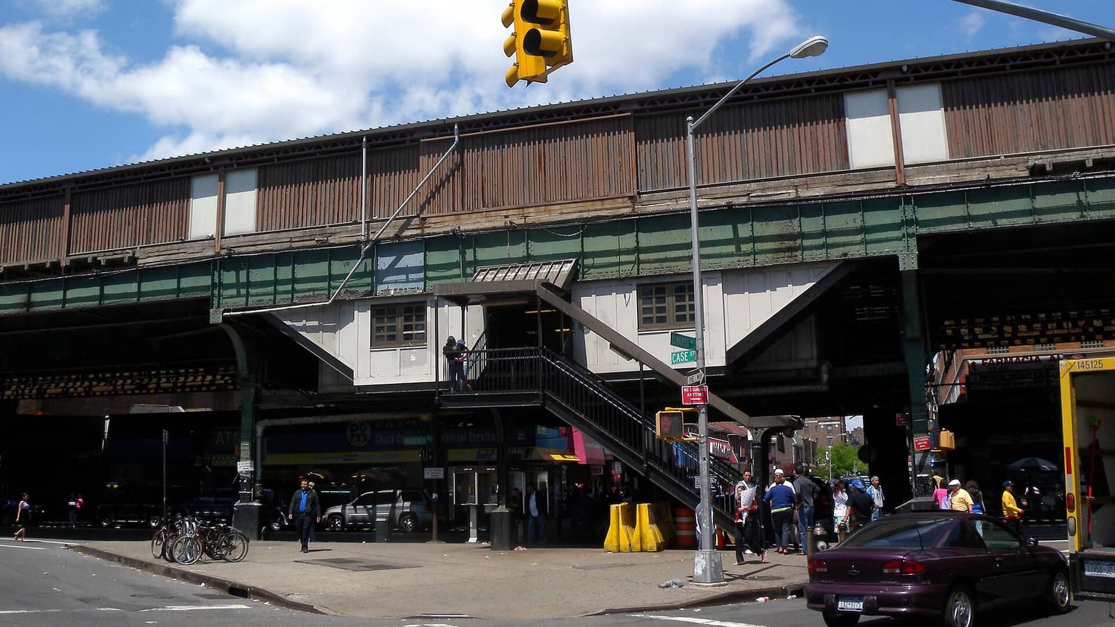 A raised subway station of the Flushing Line over a street corner, with brown wooden siding, a green steel structure, a stairway down to the sidewalk, a yellow traffic light, parked bicycles, shop signs under the tracks and people walking at street level