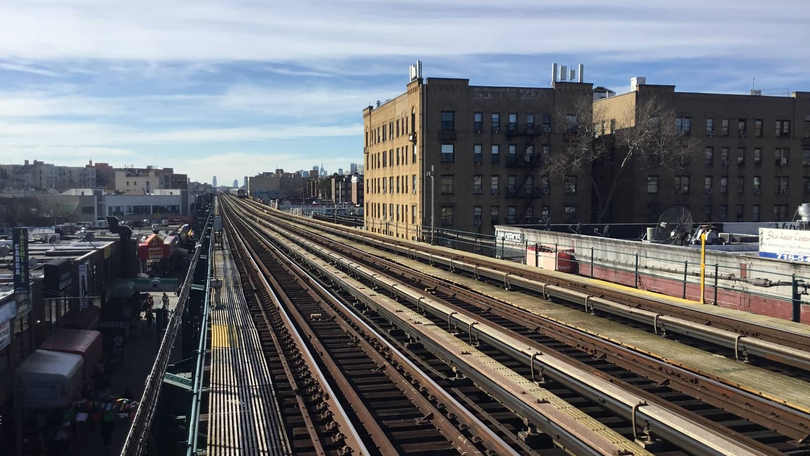 View along four raised subway tracks toward the Manhattan skyline, with yellow-brick apartment buildings at right and low shops and a busy sidewalk under the tracks at left