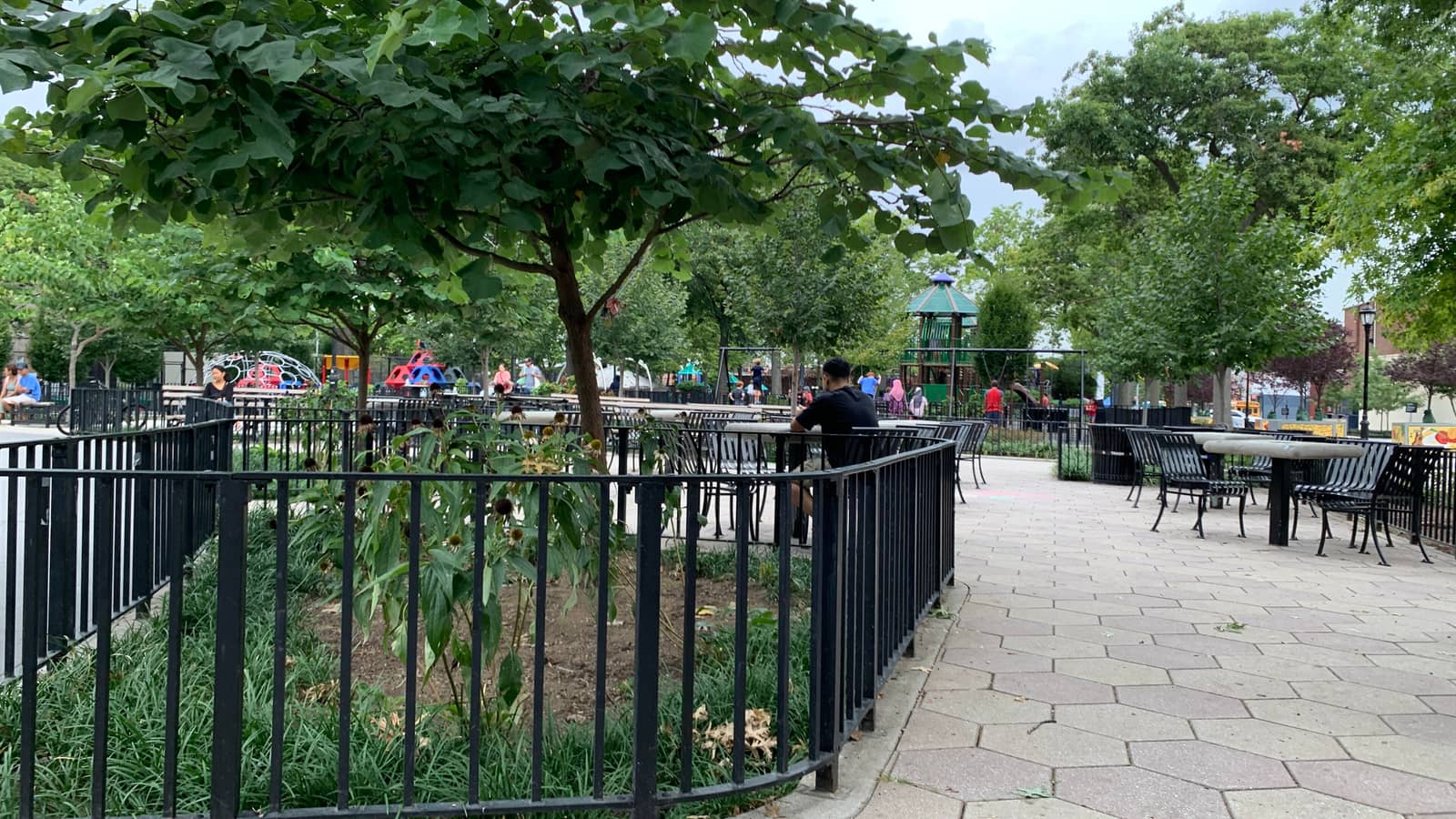 A park with a fenced flower bed and trees, a paved path with metal tables and chairs, and a playground with a green gazebo and climbing equipment behind; a few people sit or walk in the distance