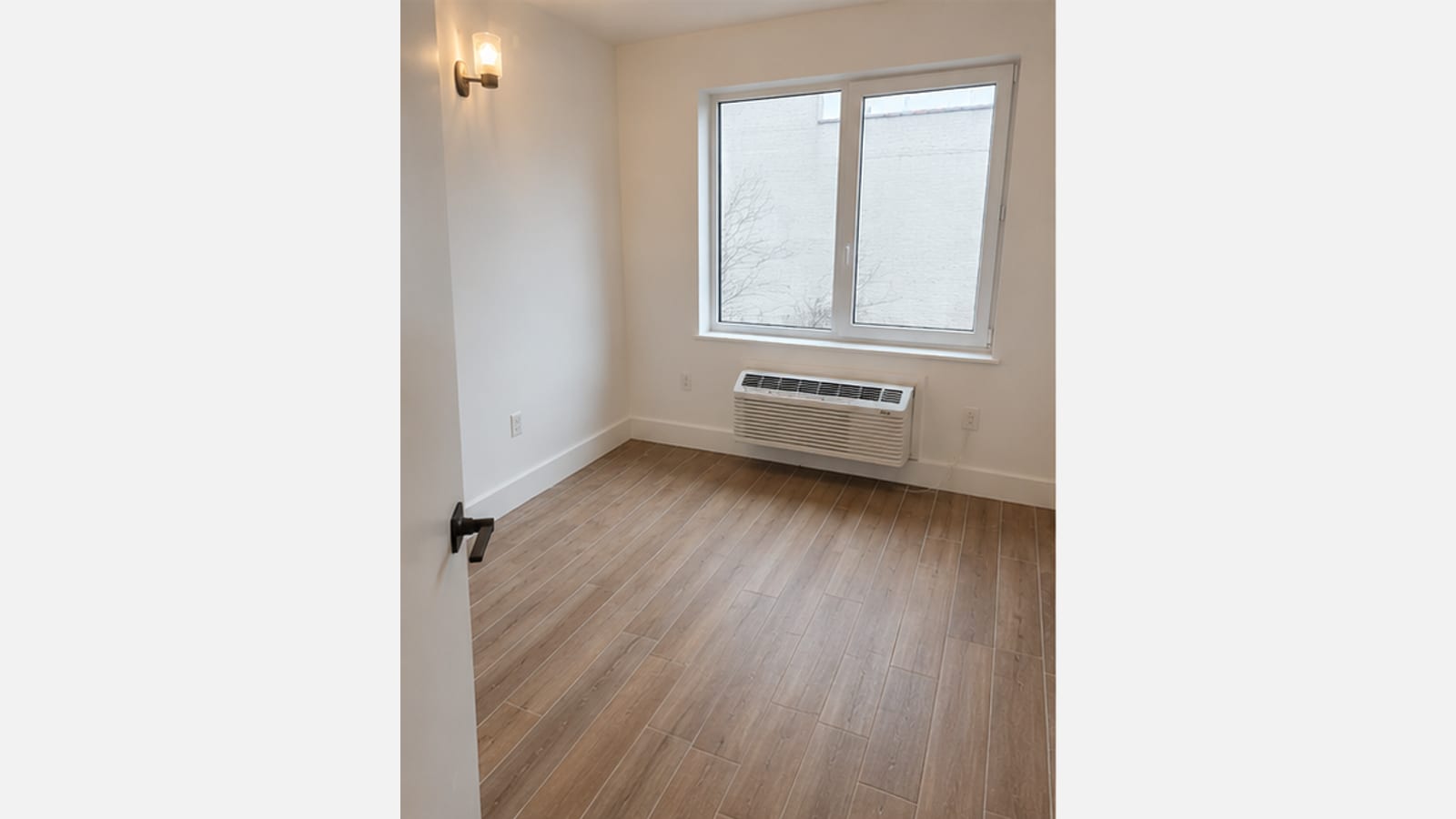 Photograph of an empty small room seen through an open door with a black lever handle: white walls, a wall light, wood-look plank flooring, a wide two-pane window looking onto a pale wall and bare tree branches, and a white through-wall unit below the window