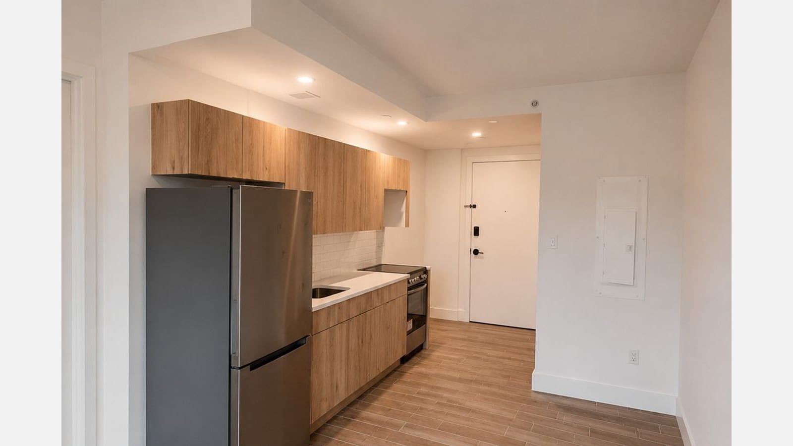 Photograph of a galley kitchen along one wall: a stainless steel refrigerator, light wood upper and lower cabinets, a white counter with a sink, white tile above it and a smooth-top electric range with an oven, with the apartment's white entry door and a wall-mounted electrical panel beyond