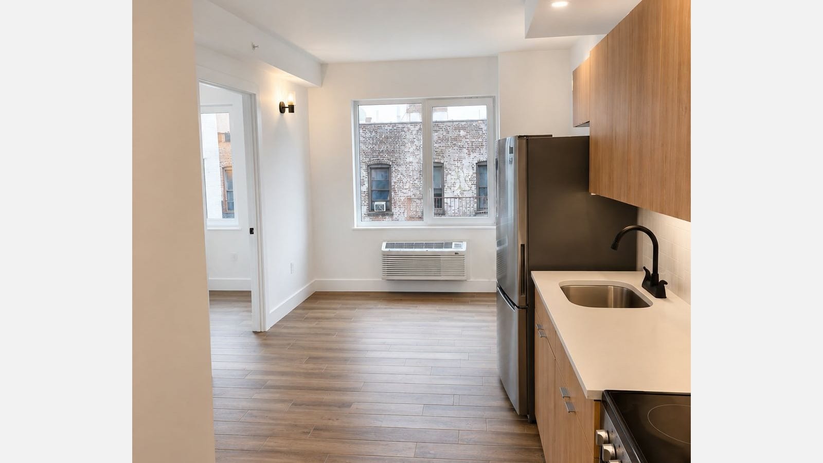 Photograph of an empty, white-walled living room with wood-look plank flooring, a wide two-pane window looking out at an older brick building with a fire escape, a through-wall unit below the window, an open door at the left into a second room, and at the right a stainless steel refrigerator beside light wood cabinets, a white counter with a black faucet and the edge of a smooth-top electric range