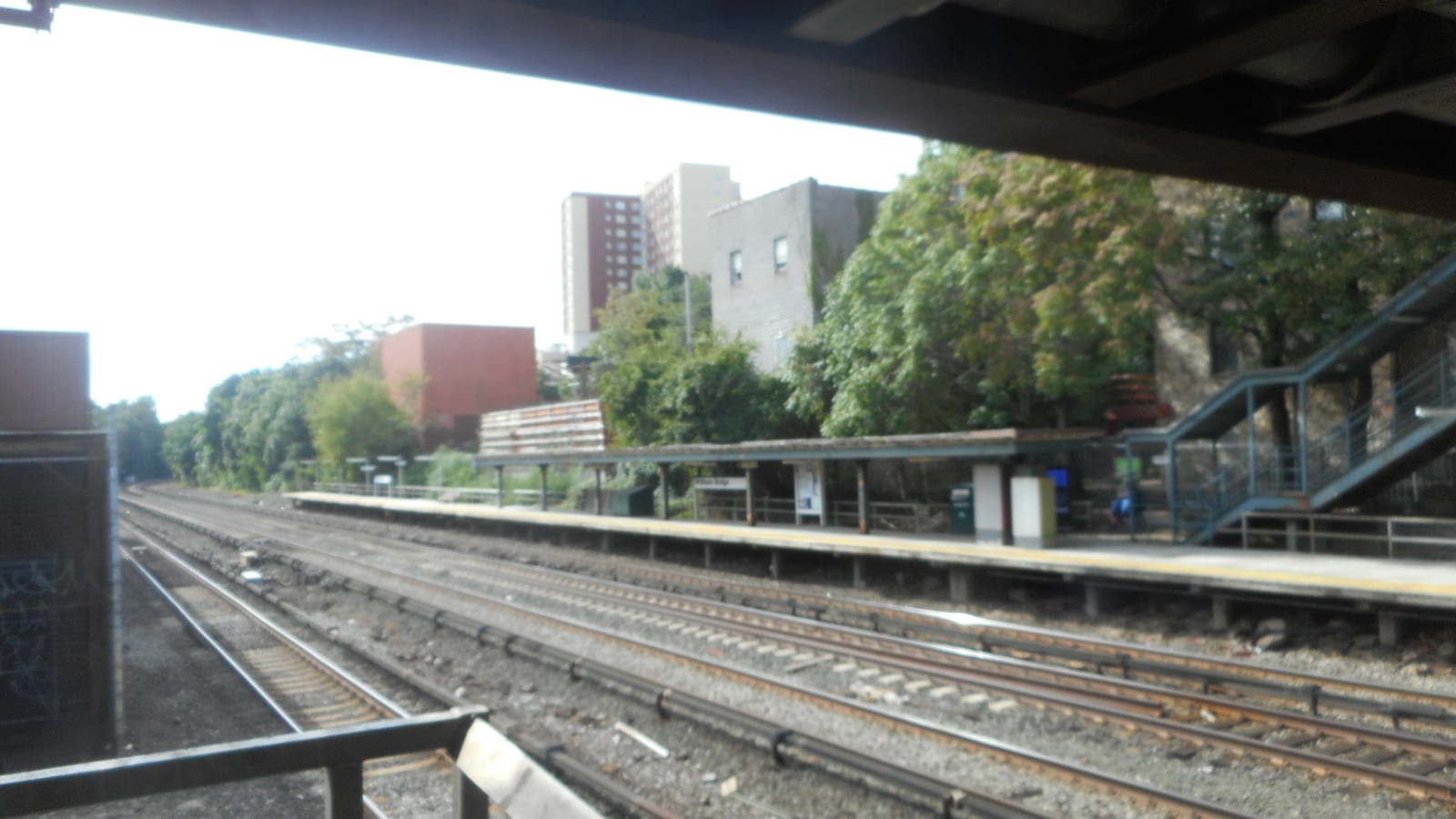 A commuter rail station seen from under a bridge: four tracks on gravel, a long platform with a low canopy and a blue staircase at right, trees and brick apartment towers behind