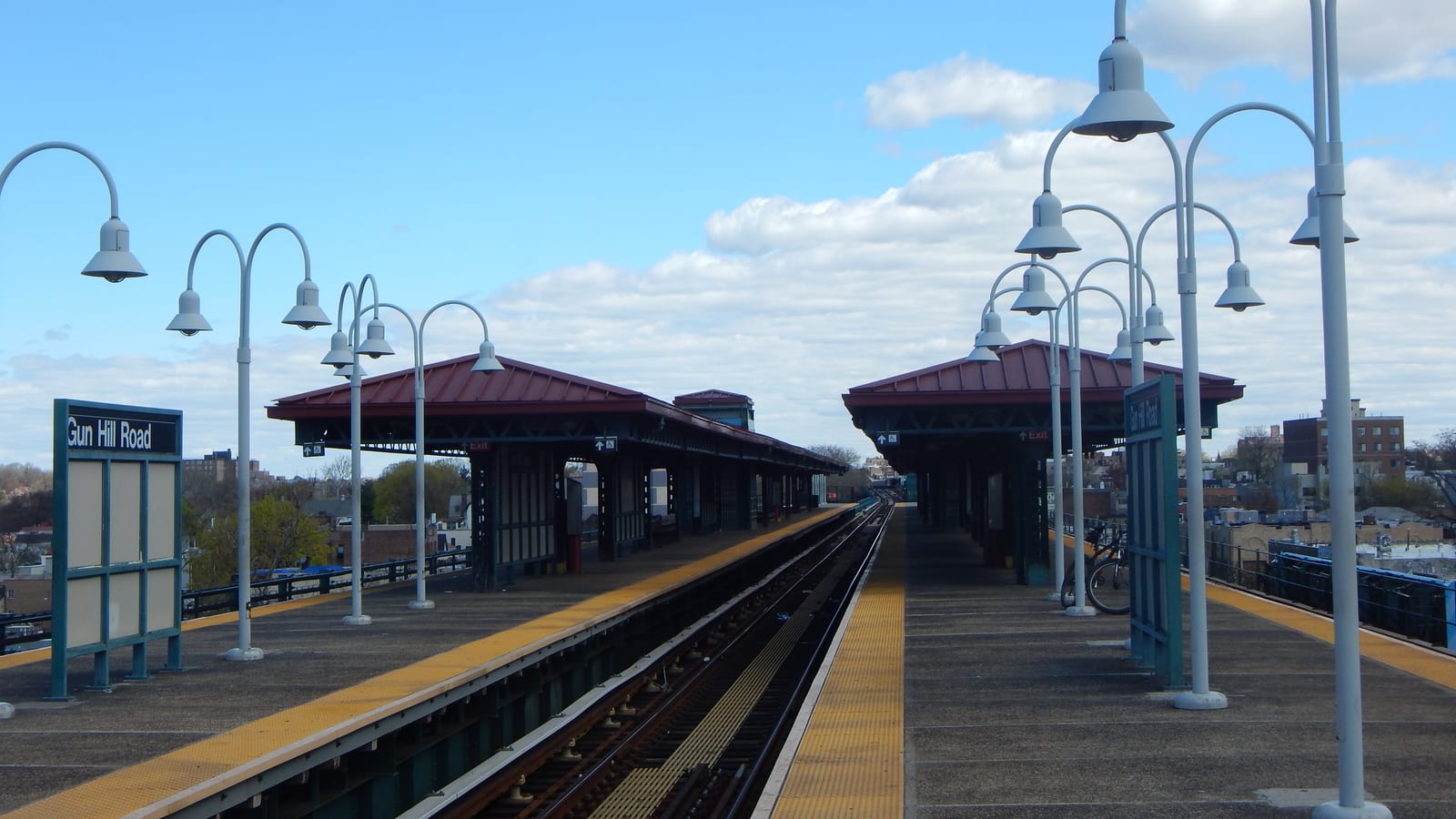 An elevated subway platform with a Gun Hill Road sign at left, tall double-headed lamp posts, red-roofed canopies over both platforms and tracks running into the distance under a partly cloudy sky