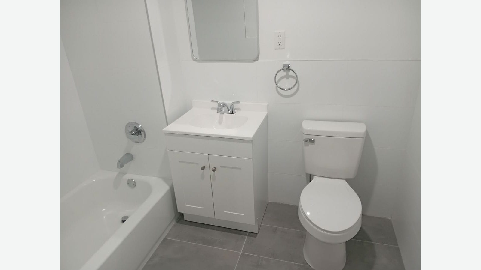 Photograph of a bathroom with white wall tile, a white tub with chrome fixtures at left, a small white vanity with a chrome faucet below a rounded mirror, a towel ring, a white toilet and large gray floor tiles