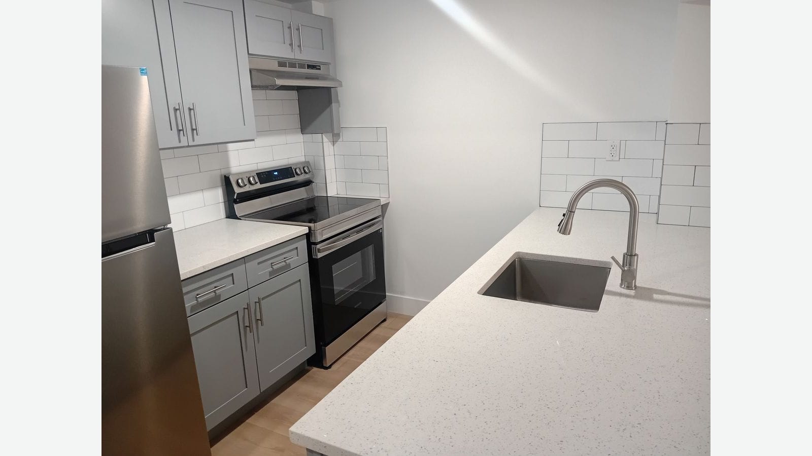 Photograph of a kitchen with a white speckled counter and stainless sink with a pull-down faucet in the foreground, gray shaker cabinets, white subway tile, a stainless electric range with a smooth top, and the side of a stainless refrigerator at left